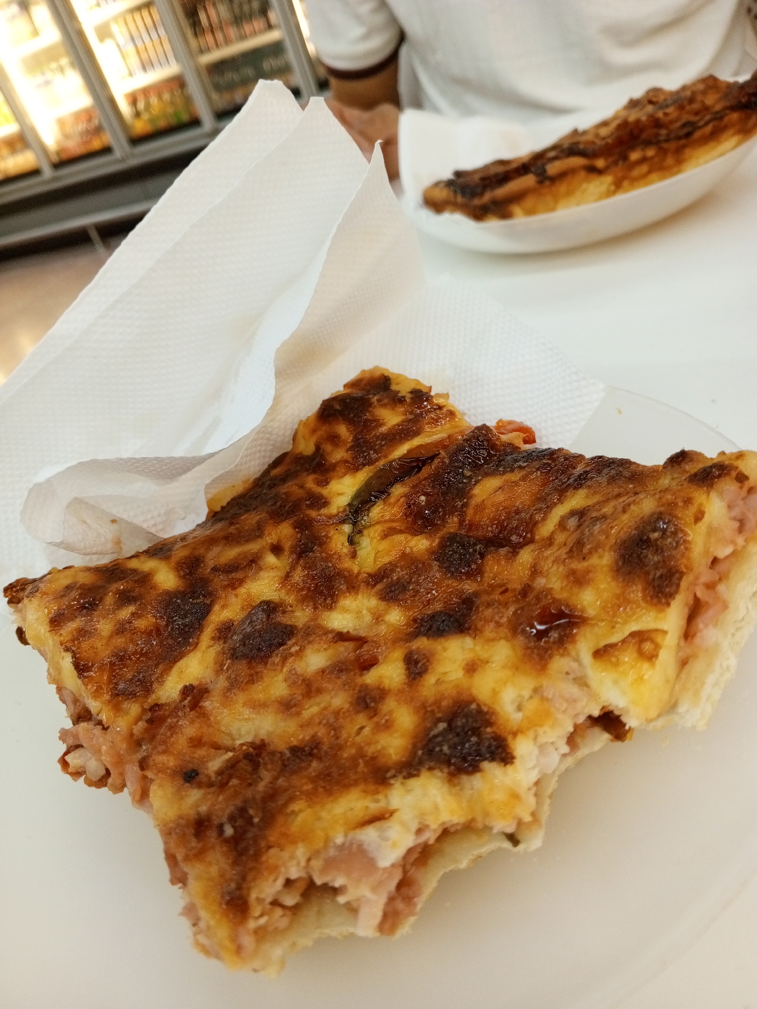 The image shows a close-up of a partially eaten piece of quiche on a white plate, accompanied by a white paper napkin. The quiche appears to have a cheese and ham filling, with a browned top. Another plate containing what appears to be the remaining portion of the quiche is visible in the background, slightly out of focus.
A person is partially visible in the upper portion of the image, wearing a white polo shirt. In the background to the left, a refrigerated display case, likely containing beverages and food items, can be seen. The picture appears to have been taken indoors, possibly in a cafe or restaurant.
While it's impossible to definitively determine the location in Porlamar, Venezuela without additional context, based on the presence of the type of food and the environment, it is likely a cafe or restaurant setting.
