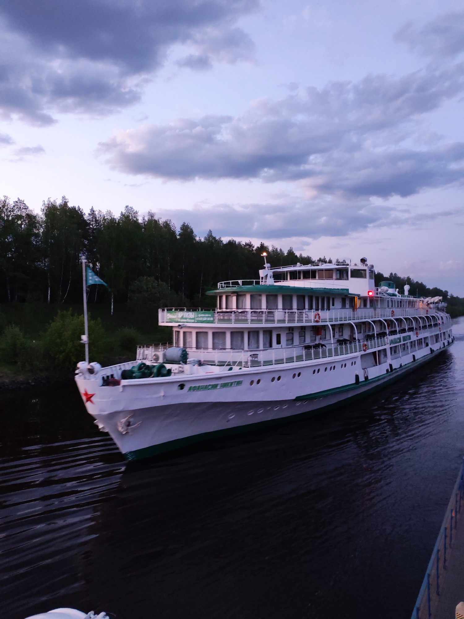 A large, white, multi-deck river cruise ship named "АФАНАСИЙ НИКИТИН" is navigating a waterway in Zaprudnya, Russia. The ship has a green stripe along the hull and green railings on the decks. A red star is visible on the bow, and a green flag is displayed. The water is dark and choppy, suggesting the ship is underway. The ship's lights are illuminated. The background includes a dense forest along the waterway and a cloudy sky. A blue guard rail is in the bottom right corner.