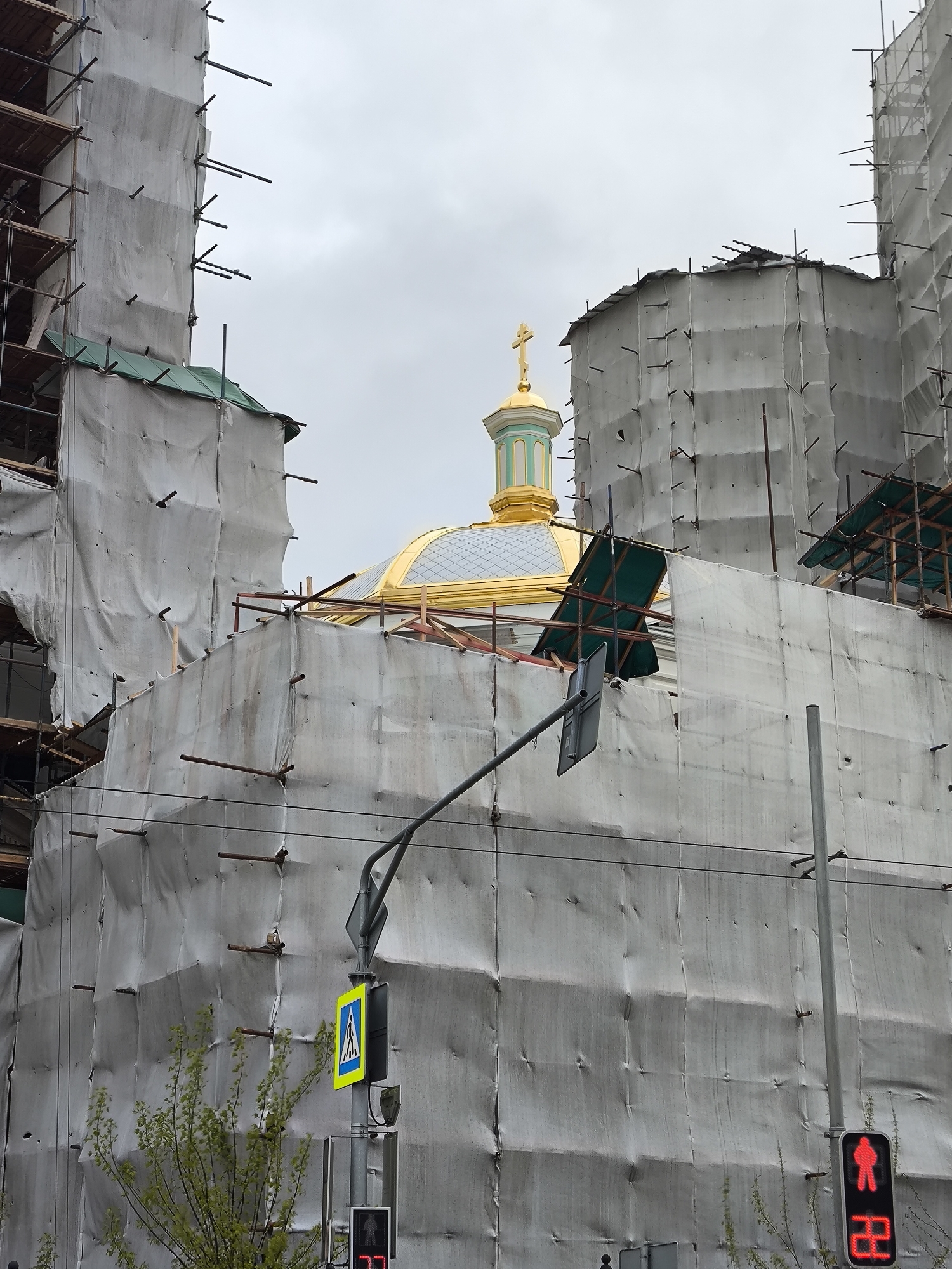 This image captures a large building, likely a church or cathedral, undergoing extensive construction or renovation in Moscow, Russia, under an overcast sky. The structure is almost entirely encased in light gray or white protective sheeting, supported by a dense network of scaffolding, with numerous metal poles visible through and around the tarps.
Emerging from the top of the covered edifice is a distinctive golden dome with a light green octagonal base, topped by a golden Orthodox cross. This architectural detail strongly suggests the building's religious purpose.
In the foreground and mid-ground, urban infrastructure elements are prominent. A metal pole supports a black traffic light signal, which displays a red "standing man" symbol and the red countdown number "22," indicating the remaining time for pedestrians. Above this, a yellow and blue square sign with a white walking person symbol clearly marks a pedestrian crossing. Thin black wires or cables traverse the upper portion of the frame. Towards the bottom left, a bush or small tree with vibrant green foliage partially obscures the base of the covered building and another small, red traffic light signal.
The scene conveys an active construction or restoration project in an urban environment, with functional street signs and signals indicating the presence of a public thoroughfare. No people are visible.