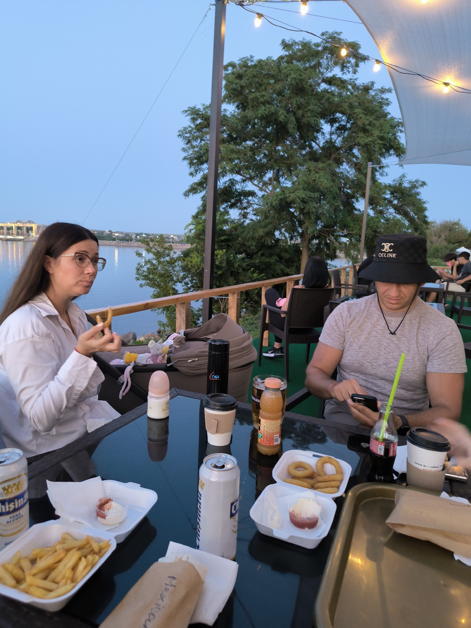 In Ustia, Moldova, on a terrace overlooking a body of water, a woman with long brown hair and glasses is eating a piece of food. She is wearing a white shirt. In front of her on a glass table are French fries, a can of soda, and a baby bottle. To her right, a man wearing a black bucket hat with the logo "CELINE" is looking at his phone. He is wearing a gray t-shirt and has a green straw in a bottle of Coca-Cola next to him. There are other people and tables in the background, suggesting a restaurant or outdoor dining area. The sky is a pale blue, and string lights are visible overhead.