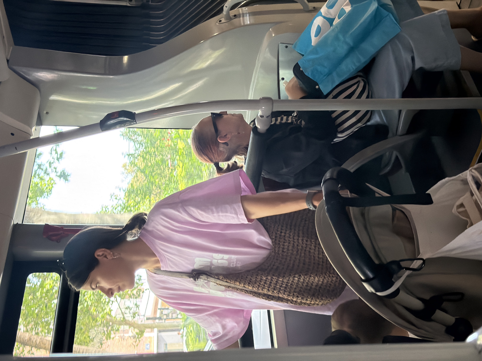 An indoor scene on a bus shows two women and a baby stroller. The woman on the left, facing left and out a window, wears a pink t-shirt and a woven brown bag. The woman on the right, facing left and wearing sunglasses, is seated with two blue shopping bags. A baby stroller is visible in the foreground. The background shows a window with trees and buildings visible outside.