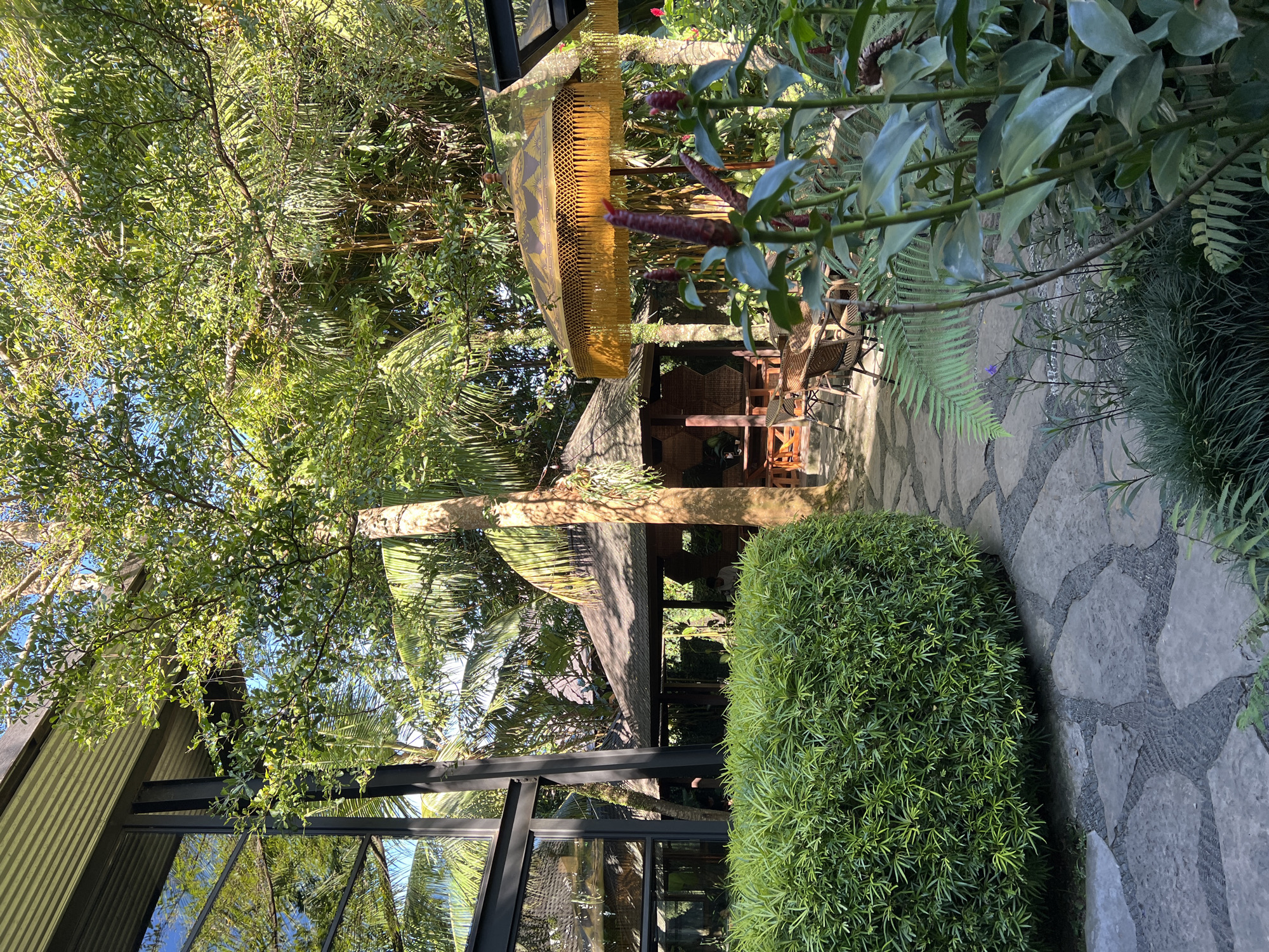 The image captures a garden scene in Sukawati, Indonesia, with a mix of natural and architectural elements. A small structure with a thatched roof and a decorative golden umbrella is visible, likely a gazebo or seating area. The structure is surrounded by lush tropical vegetation, including trees, bushes, and flowering plants. A stone pathway winds through the garden. Part of a modern building with large glass windows is visible on the left side of the image. The reflection on the windows shows the sky, creating an interplay between the natural and built environments.