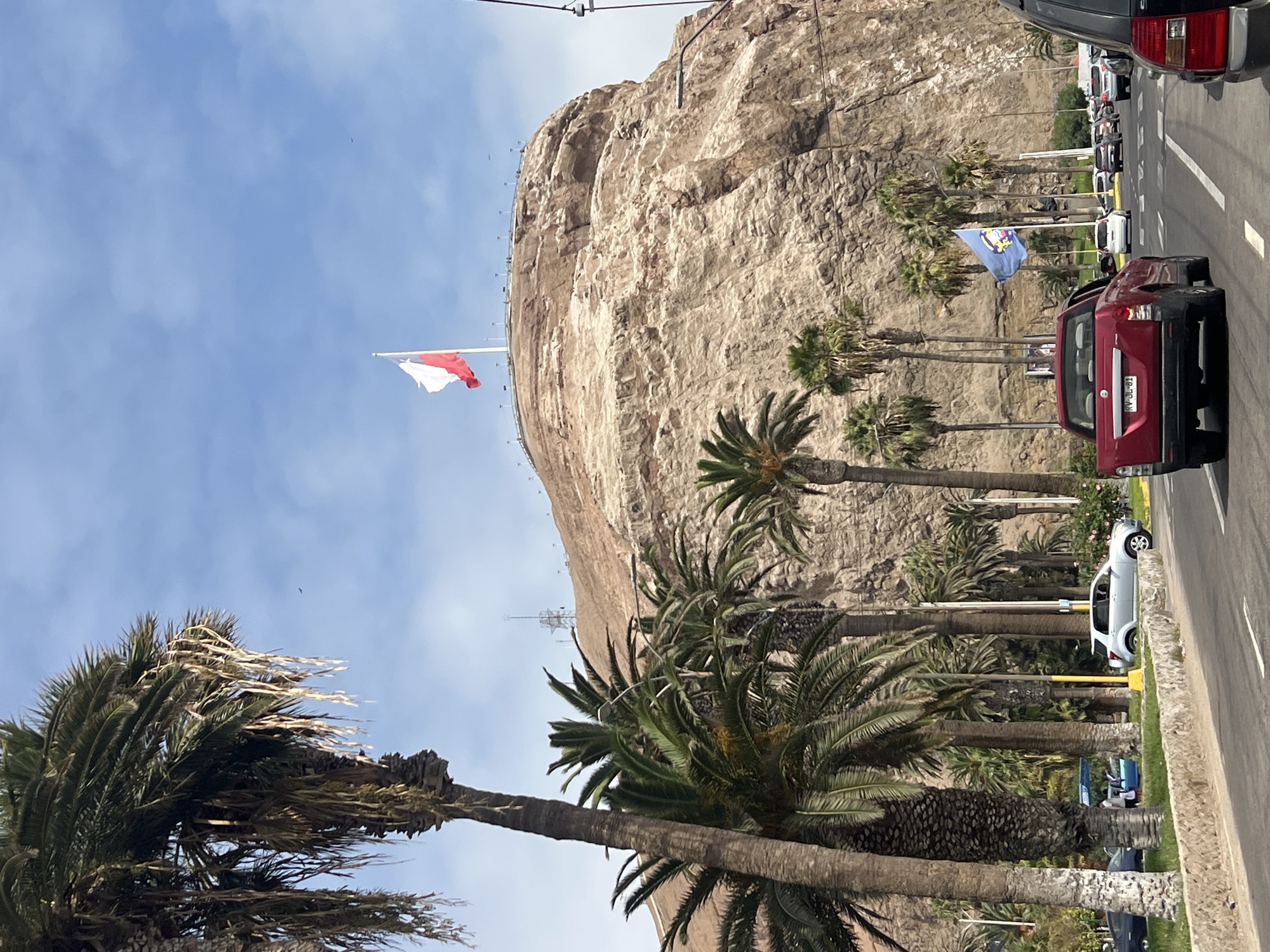The image shows a coastal street in Arica, Chile, lined with palm trees and cars. A large rock formation dominates the background, flying the Chilean flag. There are cars on the road, and the environment appears to be urban and coastal.
