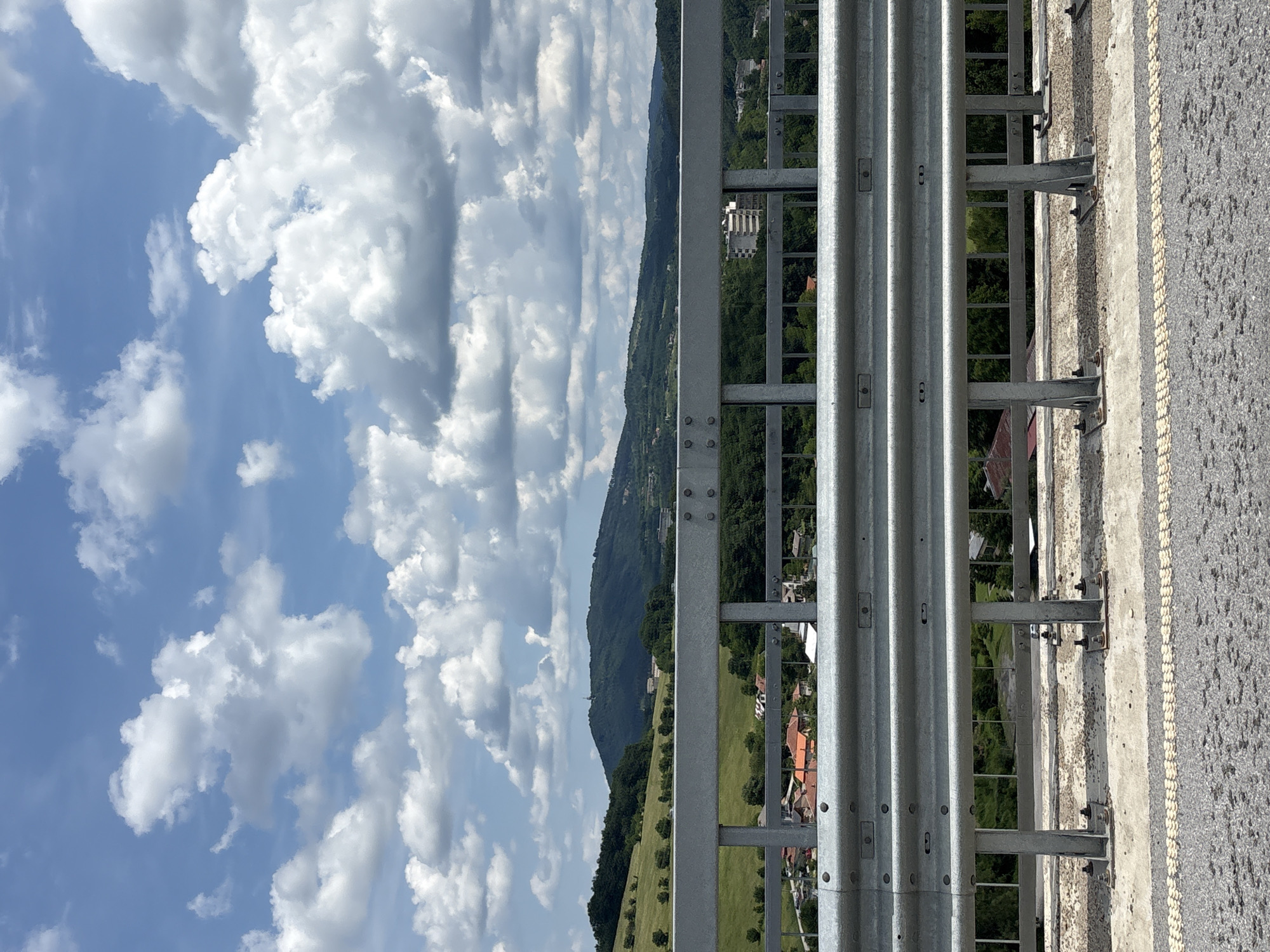 The image shows a view of a hilly landscape from a bridge in Gabrovo, Bulgaria. In the foreground, a metal guardrail with two horizontal beams is prominent, bolted to vertical posts. The road surface in front of the guardrail is asphalt with a textured yellow and white line. Behind the guardrail, a valley unfolds with a village comprising red-roofed houses and scattered trees. Further in the distance, a large green hill dominates the background, with a few buildings visible on its slopes and summit. The sky is a bright blue, filled with fluffy white and grey clouds. The scene captures a typical summer day in a rural setting.