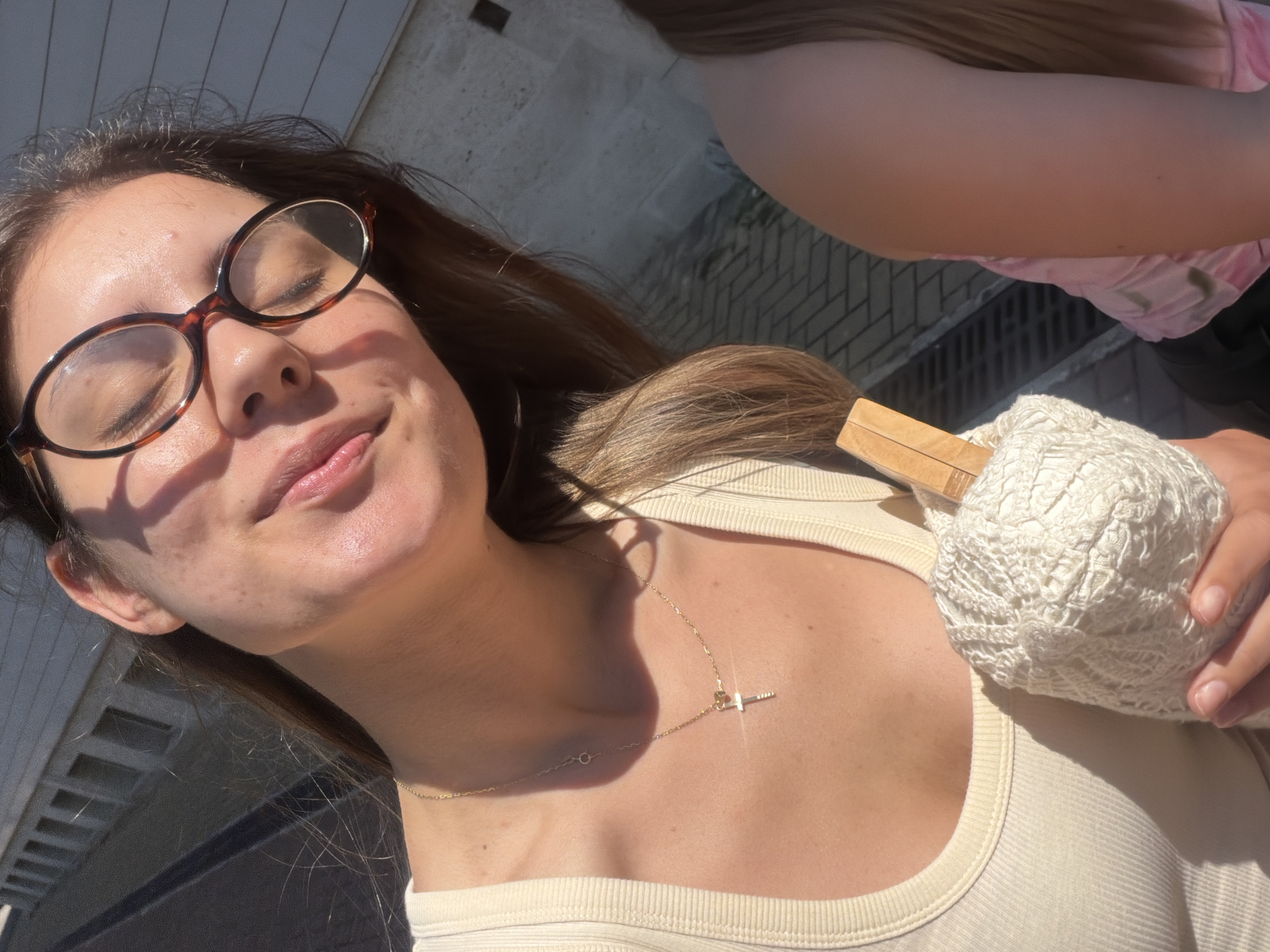 A young woman with long brown hair, wearing glasses and a cream-colored ribbed tank top, is captured in a close-up, selfie-style shot. She is holding a ball of white yarn with a wooden stick protruding from it. Her eyes are closed, and she has a gentle smile on her face, with sunlight highlighting her features. A thin gold chain with a cross pendant rests on her chest. In the background, out of focus, is another person with long brown hair wearing a pink top. The environment appears to be outdoors, possibly a street or promenade, with a building and paved ground visible. The location is identified as Burgas, Bulgaria.