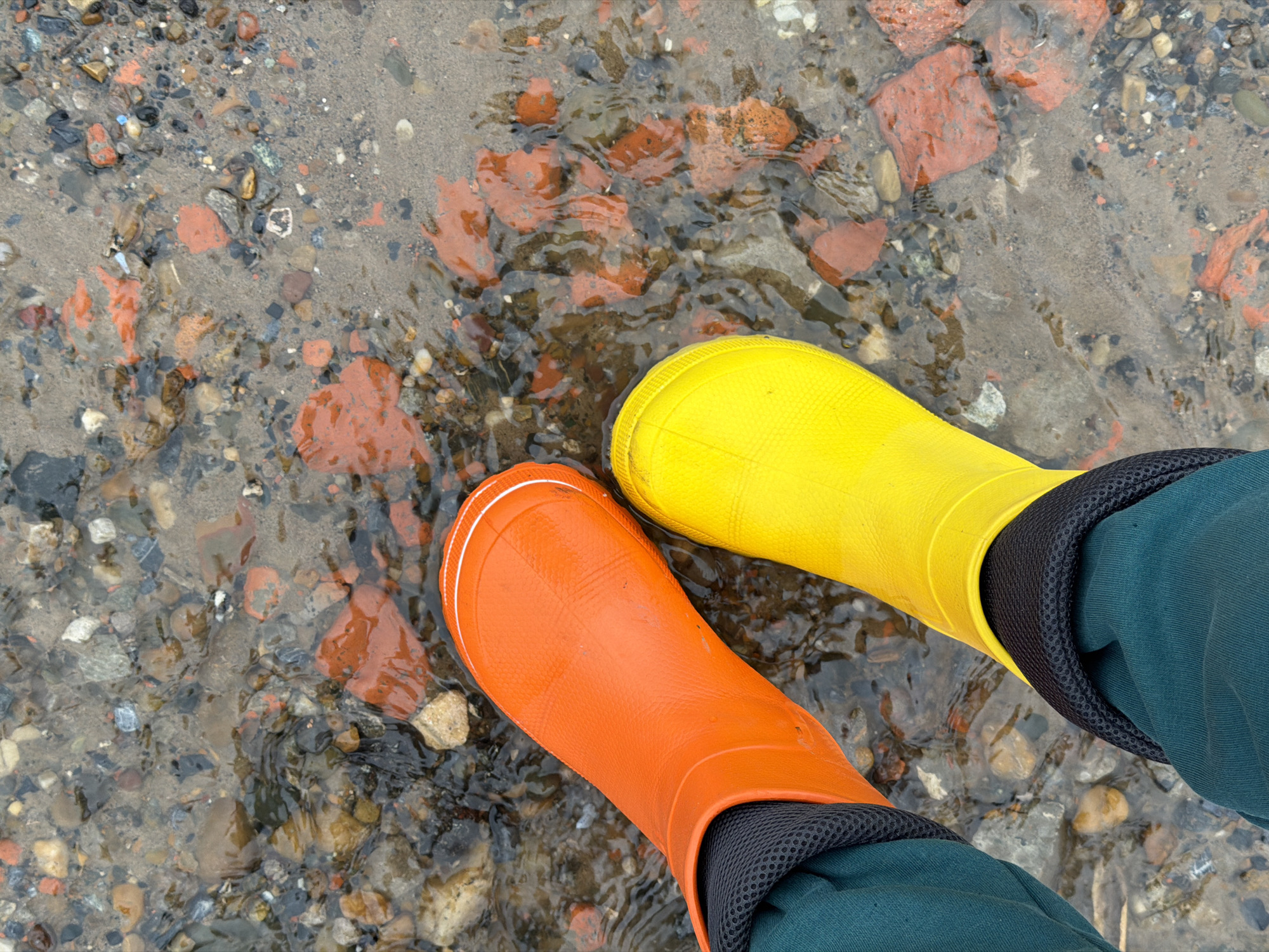 The image shows a pair of feet wearing mismatched rubber boots standing in shallow water. The left foot wears an orange boot, while the right foot wears a yellow boot. Both boots have black trim at the top, where they meet dark green pants. The feet are standing in water covering a mixture of stones, pebbles, sand, and brick fragments. The location is described as Chaikovsky, Russia.