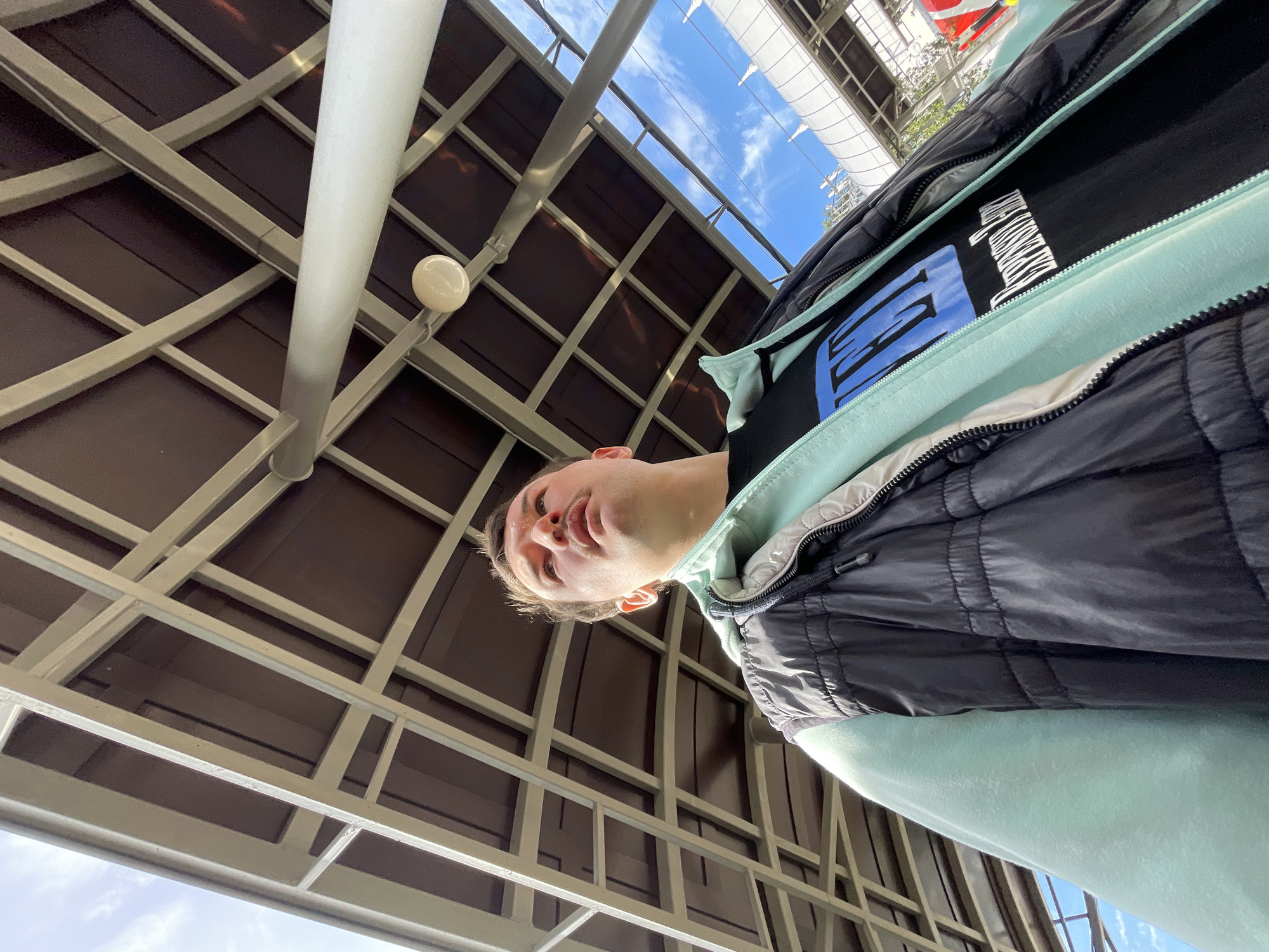 An upward-angle shot of a person under a canopy with a grid-like structure in Sochi, Russia. The canopy is made of metal supports with brown panels in between, and a vertical pole runs through it. The sky with light clouds can be seen through the canopy's edges. The person is wearing a black puffer jacket with a light blue hoodie underneath.