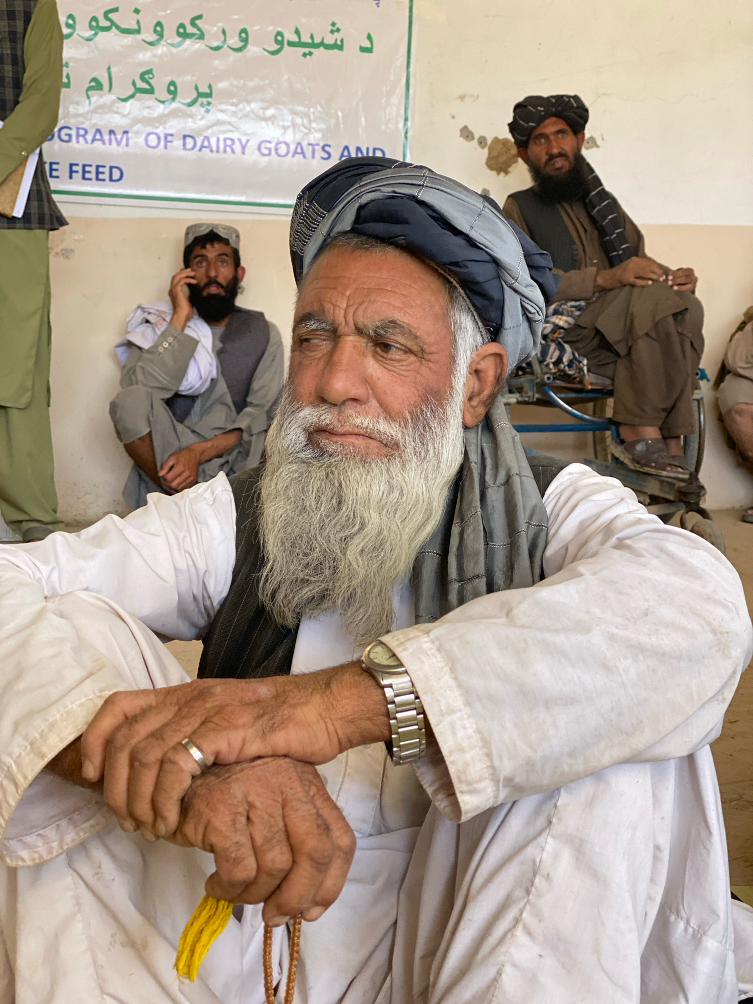 The image shows a close-up of an older man with a long white beard and a grey turban, seated in the foreground. He is wearing a white shirt and a dark vest. His hands are clasped in his lap, and he is holding prayer beads and a yellow tassel. A silver ring is visible on his left ring finger. In the background, other men are seated. One man, in the mid-ground, is wearing a grey outfit and a white cloth wrapped around his head. He is holding a phone to his ear. Another man in the background wears a dark brown outfit and a black turban. A banner with text in Pashto and English is visible behind the men, with the words "PROGRAM OF DAIRY GOATS AND" and "FEED" legible. The environment appears to be indoors with a plain, light-colored wall behind the banner. The lighting suggests it is daytime. The scene is likely in Pasab, Afghanistan, given the Pashto text on the banner and the context provided.