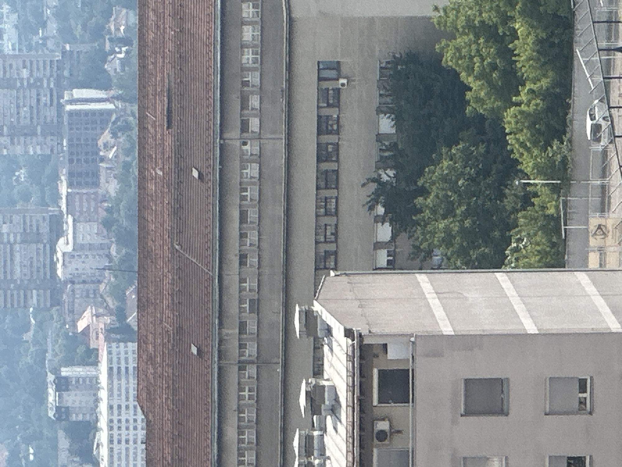 The image captures a section of a city, likely Belgrade, Serbia, with several buildings and greenery. In the foreground, a multi-story concrete building with a light grey facade features several windows, some covered with blinds. Diagonal white stripes are painted on the right side of this building's facade. Above this, another building with a reddish-brown tiled roof is visible, showing several horizontal rows of small windows.
Further in the background, a long, grey, multi-story building with a continuous row of windows runs across the middle ground. One section of this building has visible air conditioning units mounted outside. On the roof of the foreground building, several square-shaped structures, possibly ventilation shafts or chimneys with caps, are present.
Beyond these buildings, a dense collection of trees and greenery forms a backdrop. In the far distance, the hazy outlines of more buildings, characteristic of an urban skyline, are discernible. The overall scene suggests an urban environment with a mix of residential and possibly industrial or commercial structures, with nature interspersed. The lighting indicates it is daytime. No people are clearly visible in the image.