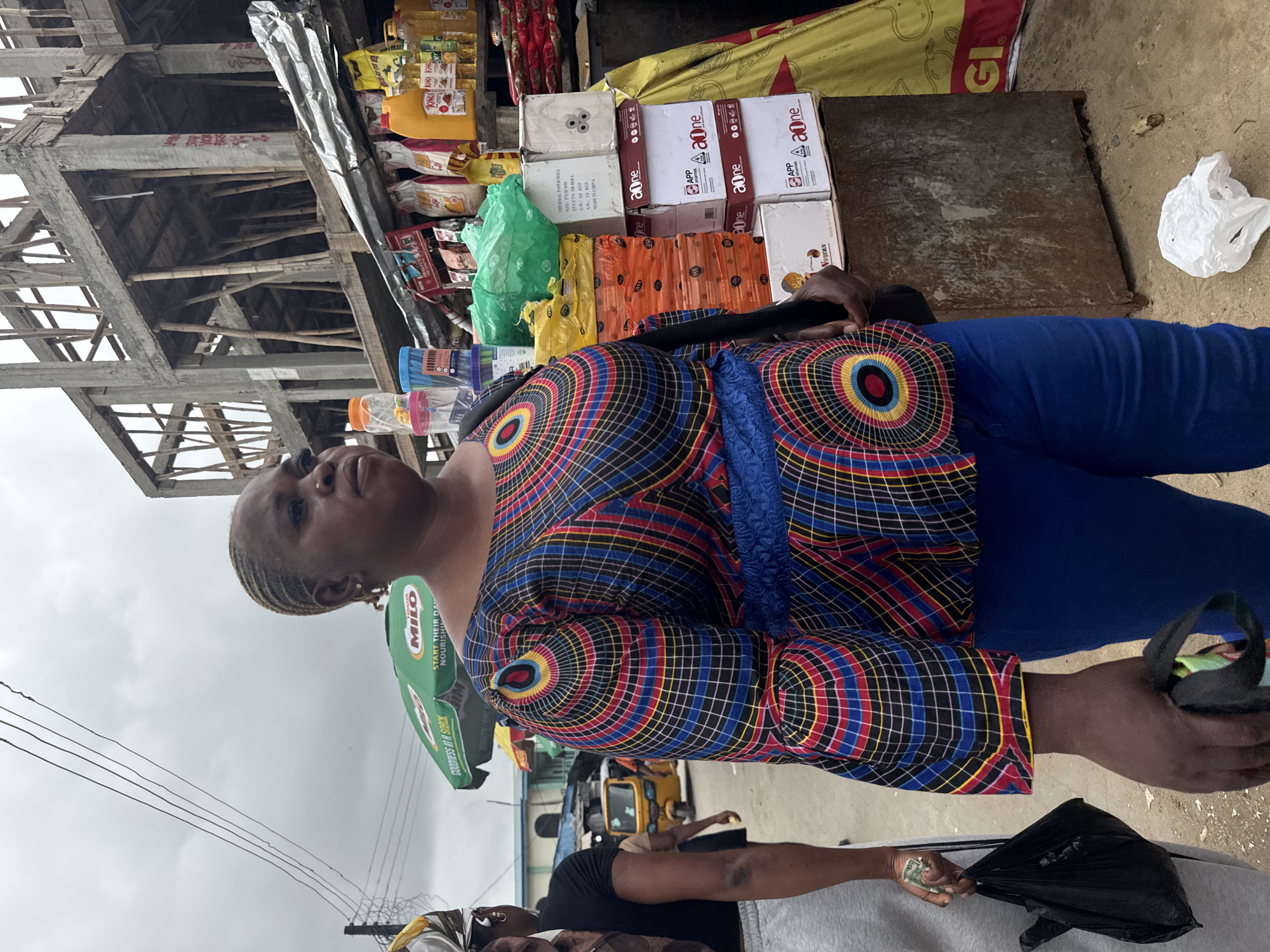 In Ado, Nigeria, a woman stands in a bustling marketplace. She wears a vibrant, patterned off-the-shoulder blouse and blue pants. Her hair is styled in braids. In her right hand, she holds a black bag. Beside her, another person, partially visible, wears grey sweatpants and a black shirt, carrying a black plastic bag in their left hand and holding a few bills of currency. The marketplace is lined with stalls displaying goods such as packaged foods, bottles of cooking oil, and stacks of paper products. Behind the stalls, a partially constructed concrete building rises, with wooden scaffolding. Overhead, power lines crisscross the cloudy sky, and a green umbrella advertising "MILO" is visible. A yellow auto-rickshaw is seen in the background.