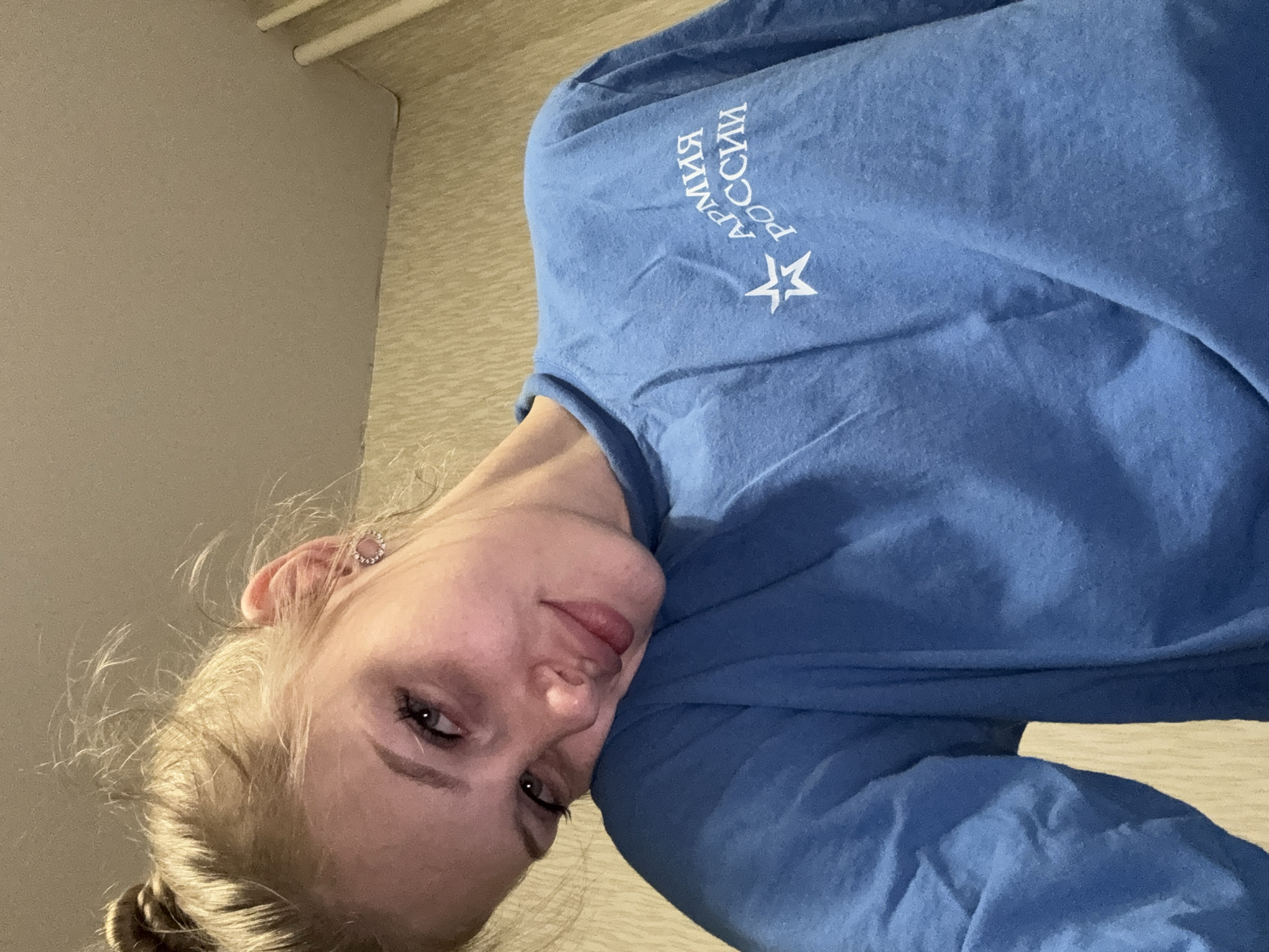A young woman with blonde hair pulled up in a bun and wearing a blue sweatshirt is shown from the chest up. Her head is tilted to her left shoulder, and she is looking towards the camera with a soft smile. She is wearing small, circular, sparkly earrings. The sweatshirt has a white emblem on the left side of the chest that includes text in Cyrillic script and a star. The background appears to be an indoor setting with textured wallpaper and a light-colored ceiling. The image was taken in Kondratovo, Russia.