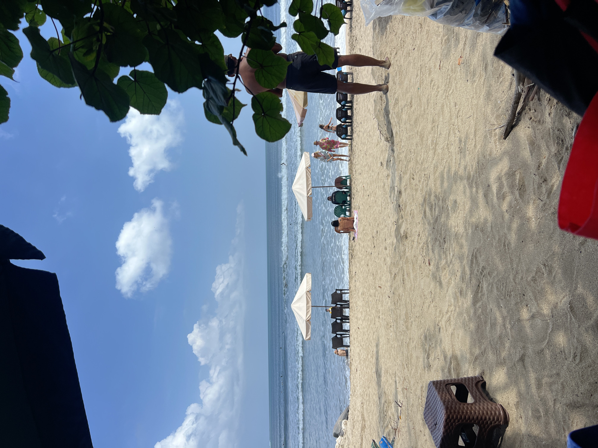This is a medium-shot, eye-level photograph taken on a sunny day at Kuta Beach in Kuta, Indonesia. The foreground is dominated by a sandy beach with some shadows cast from overhead foliage, and a brown plastic stool is visible on the left. In the mid-ground, several beige beach umbrellas are set up, with rows of empty dark plastic chairs beneath some of them. Several people are scattered across the beach: two individuals are seated near the water's edge under one of the umbrellas, while a group of four people, appearing to be two couples, are walking together further down the beach. A lone man stands near the center of the image, facing away from the camera. In the background, the ocean stretches out with moderate waves, and a few surfers can be seen in the distance. The sky is blue with scattered white clouds, and lush green leaves from a tree hang into the frame from the top right.