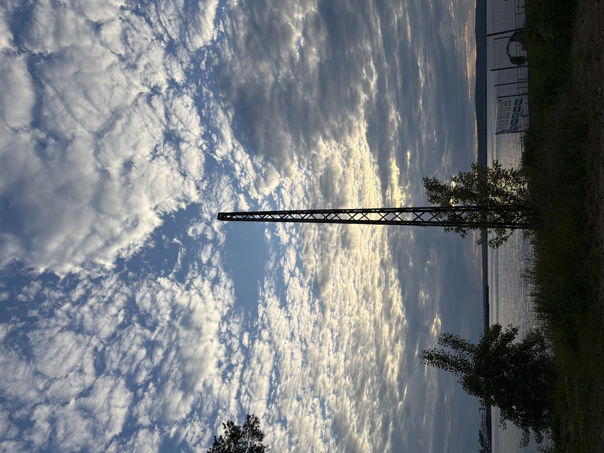The image depicts a scenic view of the Kama River in Chaikovsky, Russia, under a dramatic sky filled with scattered white clouds against a bright blue backdrop. A tall, metallic tower stands prominently in the foreground, its lattice structure silhouetted against the sky. To the left of the tower, a lush green tree with sparse foliage leans towards the water, while another smaller tree stands near the tower. The calm surface of the river reflects the soft light of the sky. In the background, a distant shoreline with rolling hills is visible, and on the water, a large ship or barge can be seen. A chain-link fence runs along the right side of the foreground, with a white sign bearing a phone number and text in Cyrillic script attached to it. The overall impression is one of natural beauty enhanced by industrial elements, captured during what appears to be either sunrise or sunset due to the soft, diffused light.