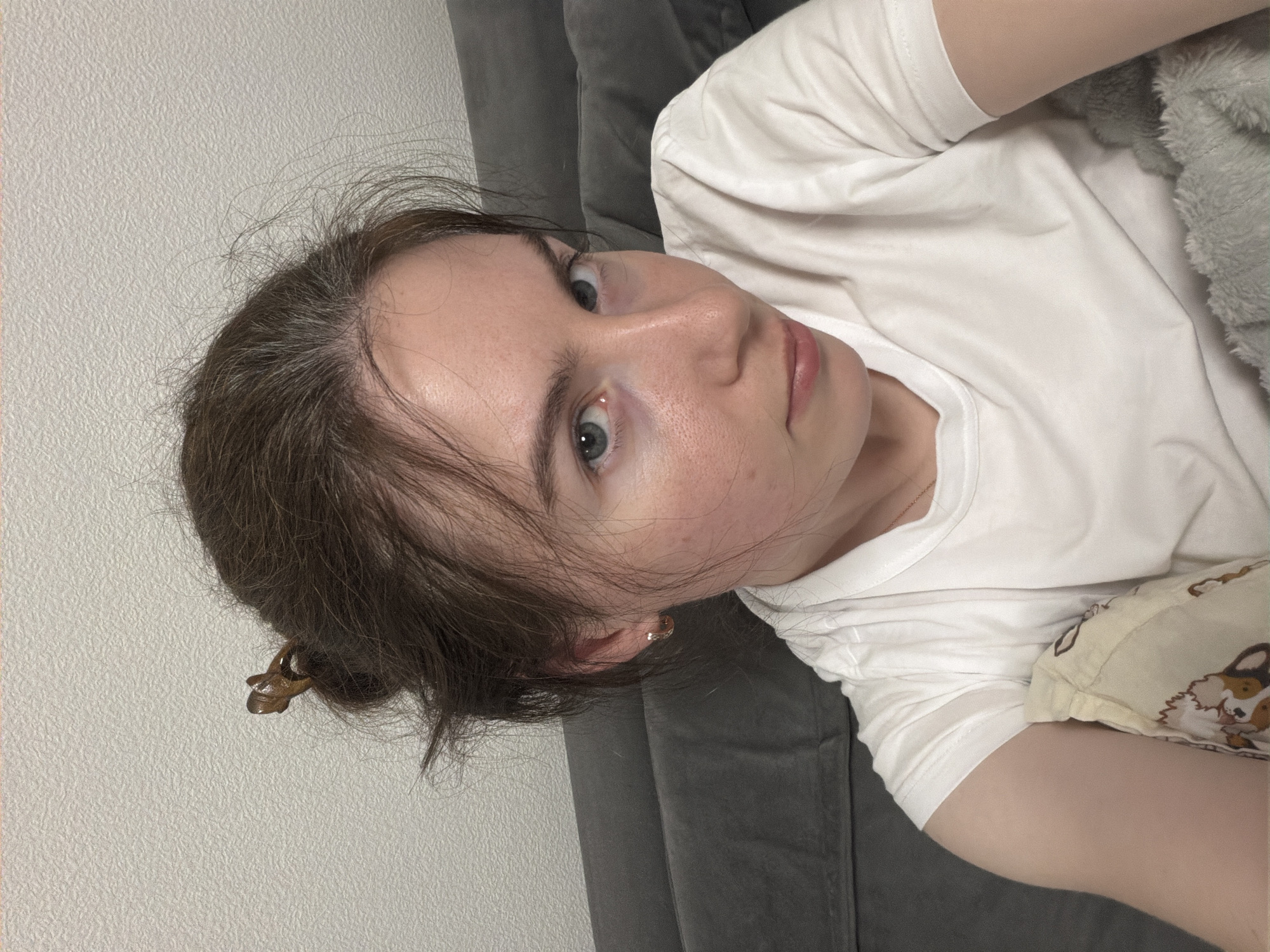 A woman lies on a gray sofa against a white wall. She wears a white t-shirt and has her brown hair pulled back with a clip. She has fair skin and blue eyes. A patterned pillow rests next to her. The location is unknown and cannot be determined from the image.