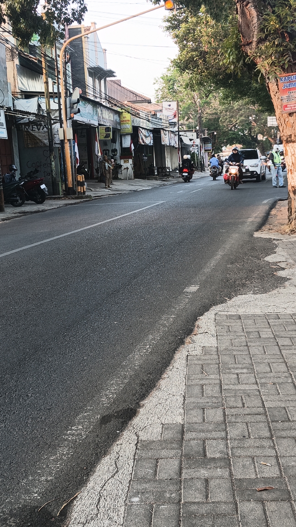 The image captures a street scene in Dawe, Indonesia, likely during daylight hours given the illumination and shadows. The foreground is dominated by an asphalt road with a white lane marker. To the right, a paved sidewalk made of rectangular gray bricks runs alongside the road. The sidewalk appears weathered, with some cracks and debris.
On the left side of the road, a row of commercial buildings lines the street. These buildings have storefronts with awnings, and some display signs and flags. Several motorcycles are parked in front of these establishments, and one is in motion on the road. A traffic light with a yellow signal is visible above the street, mounted on a pole.
Further down the street, more vehicles, including motorcycles and a white car, are traveling. Several individuals are also present, some walking or standing near the shops, and others riding motorcycles. One person is seen carrying a child. The buildings appear to be low-rise structures, typical of a town or city in Indonesia. Lush green trees overhang parts of the street, casting shadows and contributing to the urban landscape. The overall impression is of a busy, functional street within a residential or commercial area.