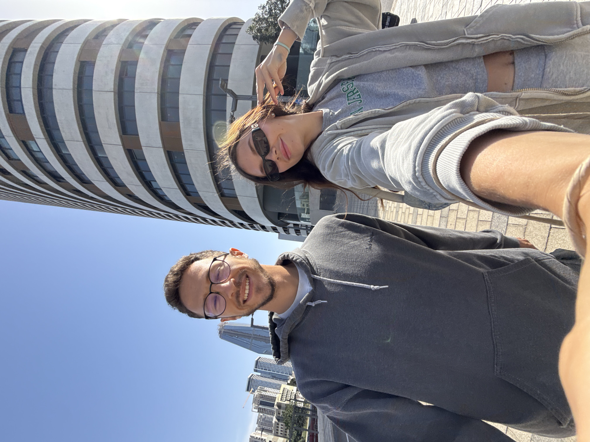 A selfie captures a man and a woman in Tel Aviv, Israel. The man, wearing round spectacles and a grey hoodie, is smiling. The woman, sporting sunglasses and a light grey zip-up jacket over a t-shirt with "Jersey" printed on it, is angled above him. The architectural backdrop includes a distinctive cylindrical building with horizontal lines and several high-rise structures. The sky is a clear blue.