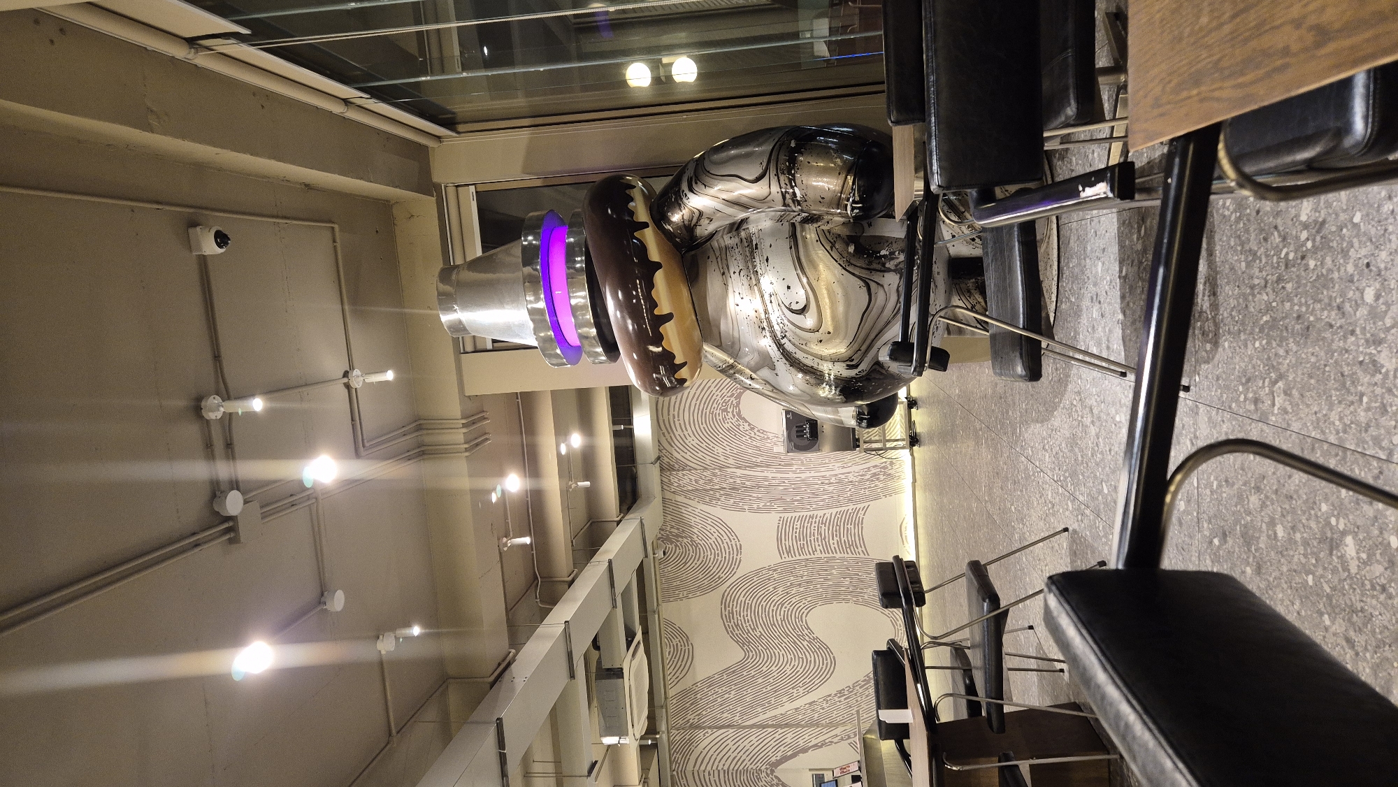 The image captures an indoor scene, likely a cafe or a retail space, featuring a large, metallic sculpture of a character wearing a donut as a hat. The sculpture has a glossy finish with swirling patterns of silver and black. Above the donut, there's a metallic cone-shaped hat with a purple glowing ring. The sculpture is positioned in the center-right of the frame. In the foreground and mid-ground, several black leather chairs with chrome legs are arranged around tables. The floor is a speckled gray material. In the background, a wall features a distinctive wavy, abstract pattern in a light color. The ceiling is visible with exposed conduits and multiple track lighting fixtures. The overall ambiance is modern and artistic. There are no people visible in the image. The image does not contain any visual cues to suggest the location is Đà Nẵng, Vietnam.