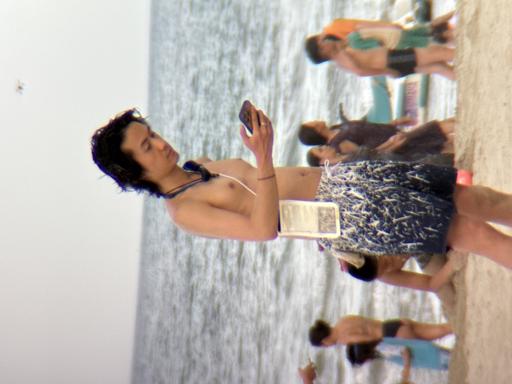 The image depicts a group of people at a beach in Da Nang, Vietnam. The primary subject is a young man lying down, holding a smartphone in his hand and looking at it. He is wearing dark swim trunks with a white and blue pattern and a thin necklace. Beside him, another person is lying down, partially visible, wearing a dark t-shirt and swim trunks. Further in the background, several other people are visible, some lying down, others standing, suggesting a relaxed day at the beach. The scene is set by the ocean, with visible waves and a sandy shoreline on the right. The lighting suggests it is daytime, likely sunny, given the bright reflections on the water. No text is visible in the image.
