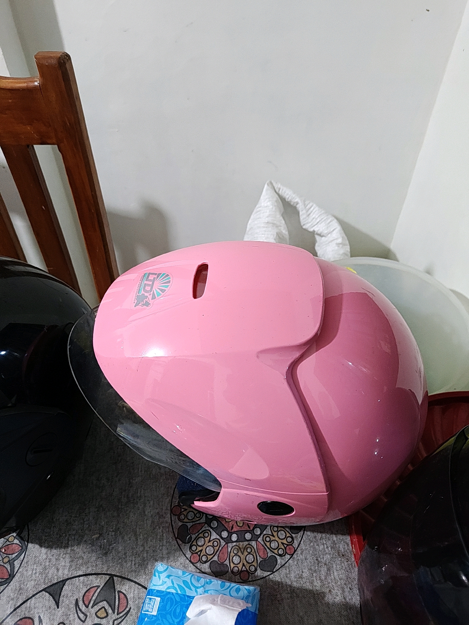 In Hutatoruan 11, Indonesia, a pink motorcycle helmet with a logo featuring a shield with a lion is placed on a textured mat. To the left of the pink helmet, a black helmet is visible, partially obscured by the pink one. In the background, part of a wooden chair and a white wall can be seen. The scene is illuminated by artificial light, casting shadows on the mat and wall.