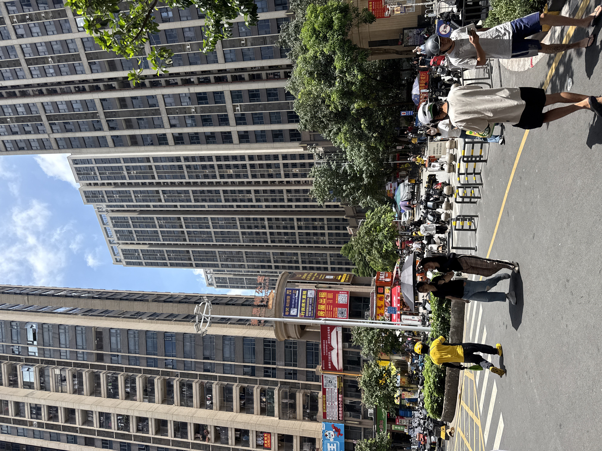 This image captures a bustling street scene in Haizhu District, China, under a bright, partly cloudy sky. Tall, modern apartment buildings dominate the skyline, their facades characterized by numerous windows and balconies. In the foreground, a street with crosswalks is visible, populated by pedestrians and a variety of parked and moving motorcycles.
Several individuals are in motion. A person in a bright yellow uniform and helmet, likely a delivery worker, is walking across the street, carrying a green bag. To their right, two women are walking together, one holding a black umbrella for shade. Further back, several people are seen walking and interacting, some wearing helmets and carrying mobile phones.
The street is lined with businesses, indicated by numerous signs and advertisements, some featuring QR codes. Green trees are interspersed with the urban architecture, providing patches of shade. The overall impression is one of a vibrant, active urban environment on a sunny day.