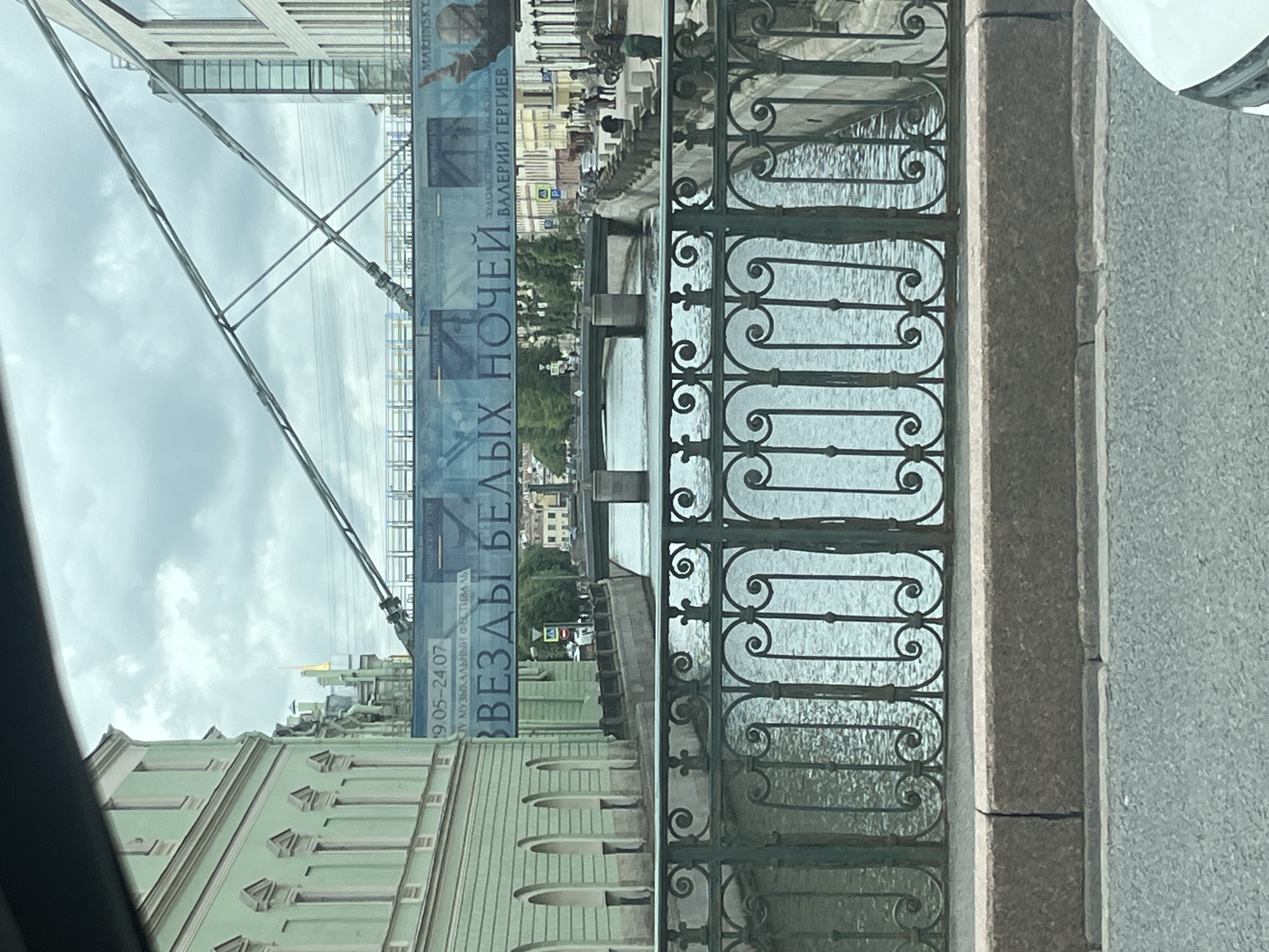 The image depicts a view from a bridge overlooking a canal in Saint Petersburg, Russia. A green wrought-iron railing runs across the foreground. Beyond the railing, the canal water is visible, reflecting the overcast sky. A pedestrian bridge spans the canal in the middle ground, with traffic and pedestrians on it. A large blue banner hangs across the pedestrian bridge, displaying text and an image of a person. On either side of the canal are buildings with traditional architecture, one with a light green facade and arched windows. The sky is cloudy, suggesting a cool or overcast day. The overall scene conveys an urban, historical setting.