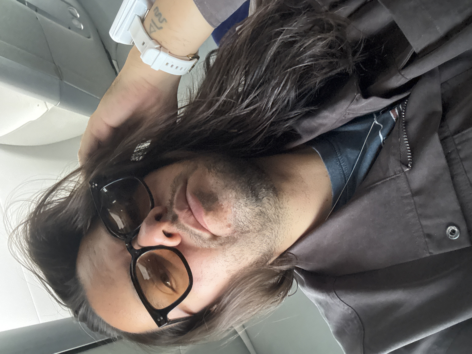 The image is a close-up selfie of a person, presented rotated 90 degrees counter-clockwise, suggesting it was taken from a low angle. The main subject is a male with long, dark hair, framing his face. He has short facial hair, including stubble on his cheeks and chin. He is wearing dark-framed sunglasses with amber-tinted lenses, which reflect some light. His attire consists of a dark grey or brown jacket, partially unzipped, revealing a dark blue or grey t-shirt underneath. On his left wrist, he wears a white digital watch, and a tattoo with indistinct symbols or script is visible on his forearm adjacent to the watch.
The background indicates an indoor environment, strongly resembling an airplane cabin, with a light grey overhead panel featuring circular vents or lights. The bright, even lighting suggests either strong artificial light or daylight filtering into the cabin. The person's expression is neutral and calm, with the sunglasses concealing his eyes. The overall scene conveys a sense of travel or transit. No legible text is present on the tattoo or watch.