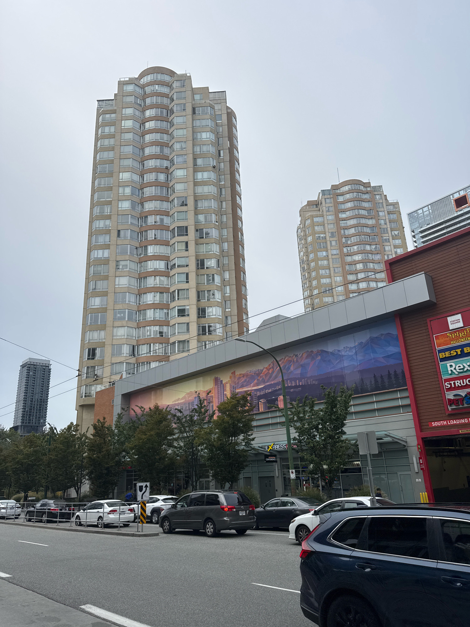 This is a street-level, eye-level, medium-shot photograph of a street scene in Burnaby, Canada, likely near a commercial or residential area. The sky is overcast.
Dominating the background are two tall, modern apartment buildings. The larger building on the left is a light-colored, multi-story structure with a curved facade and many windows. To its right, a similar, but slightly shorter, building also features a curved facade and numerous windows. In the far background, a very tall, slender, dark-colored skyscraper is visible.
In the mid-ground, a lower building with a reddish-brown facade features a large mural depicting mountains and trees. Below this mural is a section of windows, some of which reflect the sky and surrounding buildings. A street lamp with a curved arm extends into the frame from the left.
The foreground shows a street with multiple lanes of traffic. Several cars are parked or moving along the street, including a dark blue SUV in the immediate foreground, a gray minivan, a white sedan, and other vehicles further down the road. Trees line the sidewalk area. Traffic signs and utility poles are also present.
There are no clearly identifiable individuals in the scene. The overall impression is of an urban environment on a cloudy day.