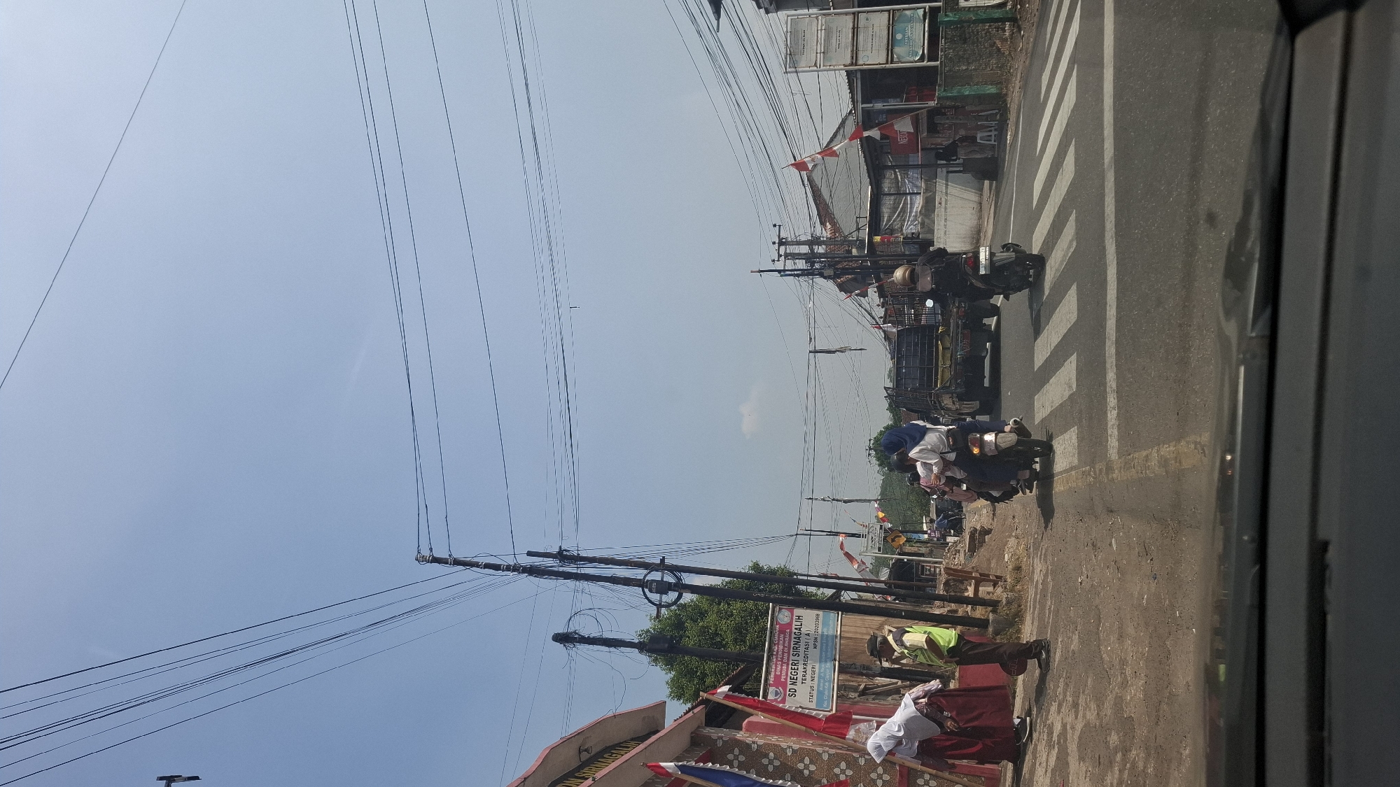 The image captures a street scene in Sirnagalih, Indonesia, on a bright, clear day. The foreground shows the interior of a vehicle, with the dashboard visible at the bottom. The road ahead is paved, with a pedestrian crossing marked by white stripes. Several individuals are present: two people are walking on the left side of the road, near a building with a sign that reads "SD NEGERI SIRNAGALIH". One of these individuals appears to be wearing a school uniform. A person in a yellow vest, possibly a school crossing guard, stands on the sidewalk. Further down the road, two motorcycles are visible. The first motorcycle, carrying two people, is crossing the pedestrian zone. The second motorcycle, also with a rider, is behind it. A large truck is stopped at the intersection, partially obscuring other elements of the scene. Various buildings and structures line the street, including what appears to be a small shop with a red "NESCAFE" sign. Overhead, a complex network of power lines crisscrosses the sky, supported by multiple utility poles. Red and white flags are visible, fluttering from some of the buildings and poles, suggesting a national celebration or event. The environment appears to be somewhat rural or a suburban area with moderate traffic activity.