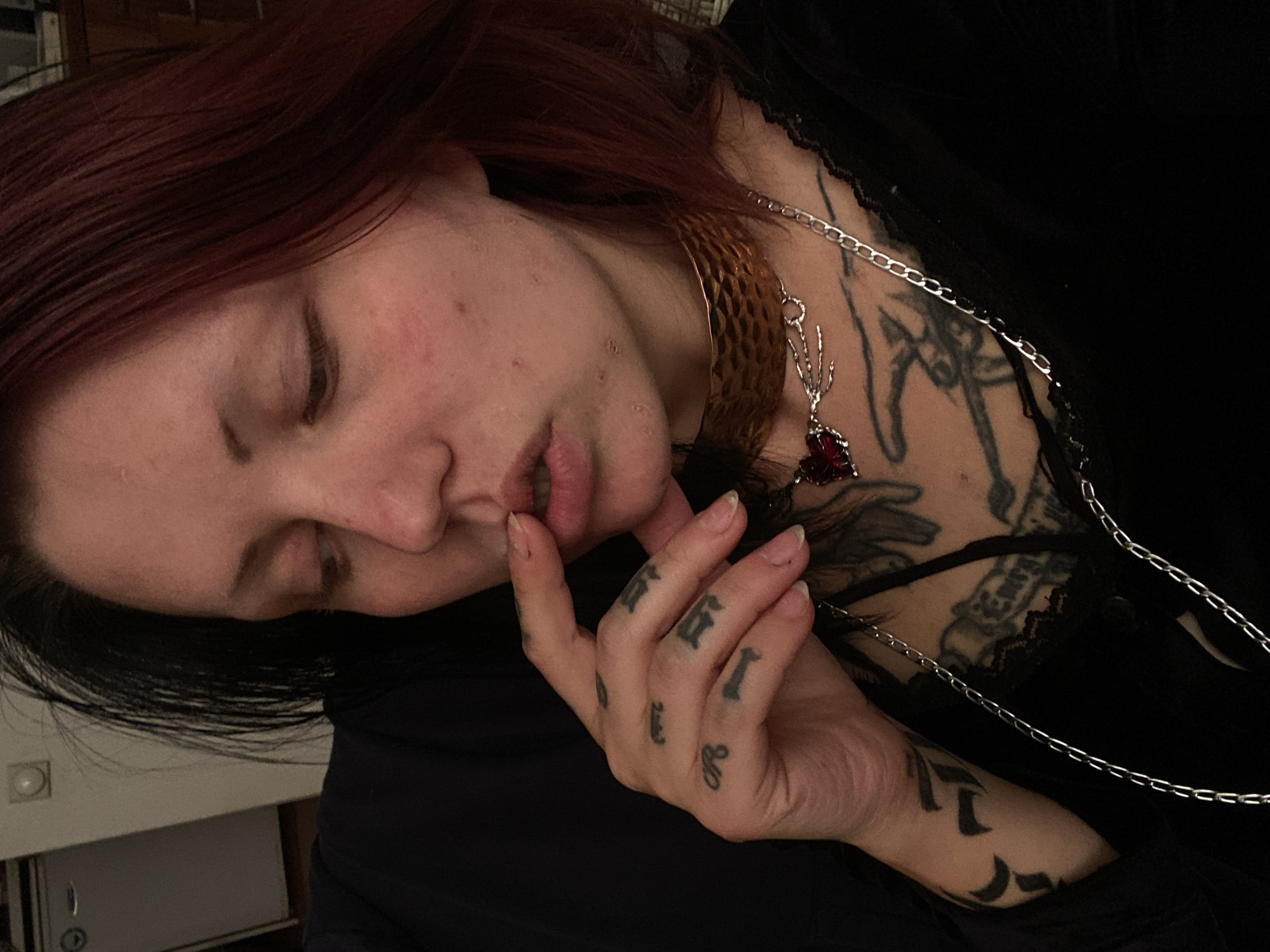 An intimate, tilted close-up shot features a person with reddish-brown hair captured in an indoor setting. The individual is wearing a black top, possibly with a lace-trimmed neckline, and is adorned with a metallic choker in a gold or bronze hue, featuring a textured surface. A silver chain necklace with a prominent red, possibly heart-shaped, pendant attached to a branching silver design is also visible.
The person's eyes are closed or heavily lidded, suggesting a moment of rest, contemplation, or introspection. Their left hand is raised, with fingers gently touching the lips. Numerous dark-inked tattoos are visible on the person, including intricate designs on the chest and collarbone area, and small symbols or letter-like characters tattooed on the fingers (e.g., 'E', 'I', 'R', and a crescent moon on the side of the palm). The fingernails are natural and unpolished.
The background is softly blurred and dimly lit, revealing faint indoor elements such as a light-colored wall with a circular fixture (possibly a light switch) and a rectangular object below it, indicating a casual home environment. The low lighting suggests the image was taken during the evening or in a moodily lit space. No specific activity is clearly depicted beyond the person's pose; the overall mood conveyed is quiet and thoughtful. No explicit text is visible beyond the tattooed symbols.
