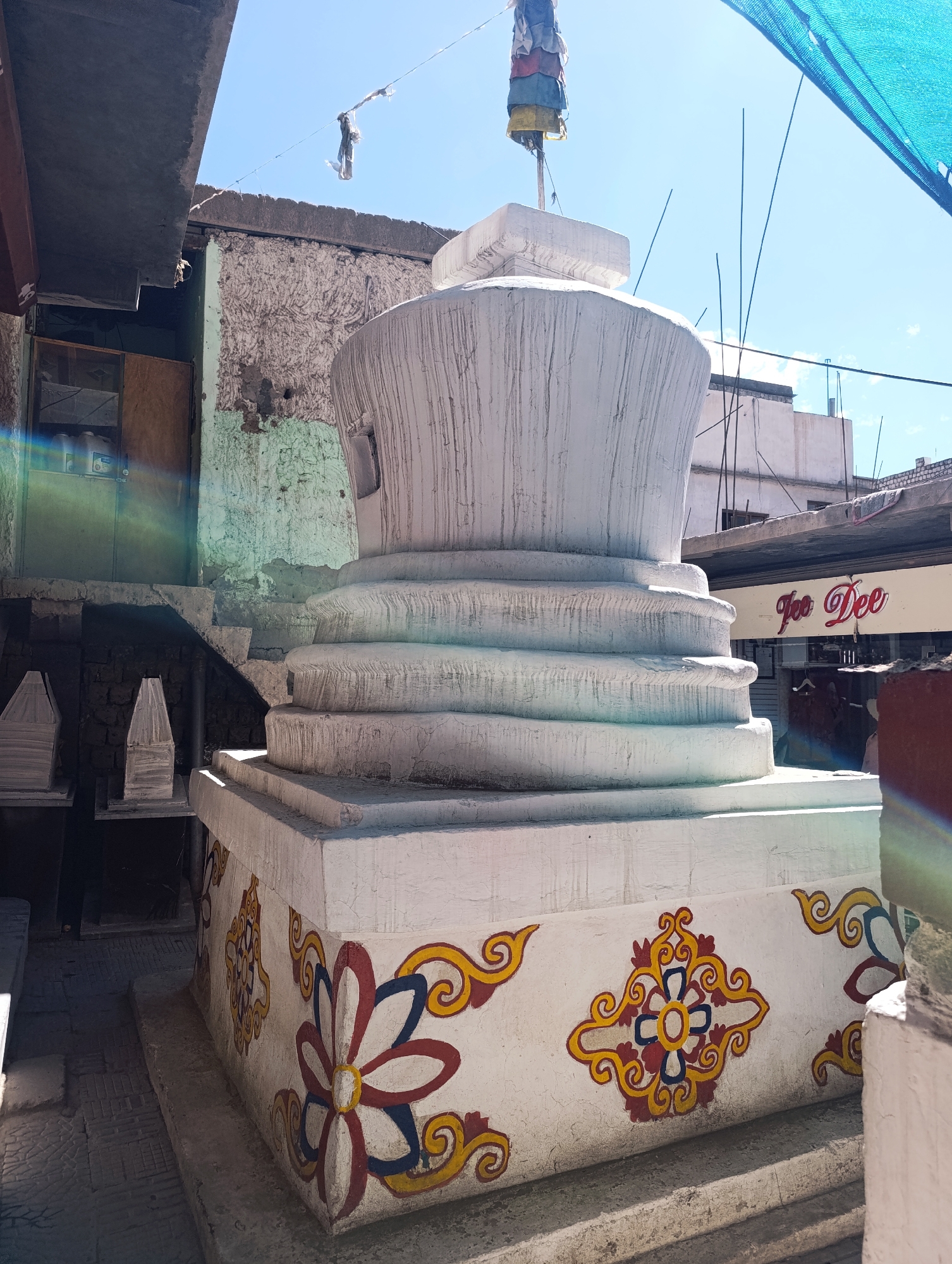 A stupa with intricate floral and scrollwork motifs painted on its base stands in the foreground. The stupa is white and appears to be made of concrete or plaster. It has multiple tiered layers leading up to a rounded dome, topped with a prayer flag. Behind the stupa, there are buildings with weathered facades and a blue tarp stretched overhead. A sign that reads "Joo Dee" in red lettering is visible on one of the buildings. The environment suggests an outdoor setting in a somewhat urban, possibly traditional, area. This image was captured in Leh, India.