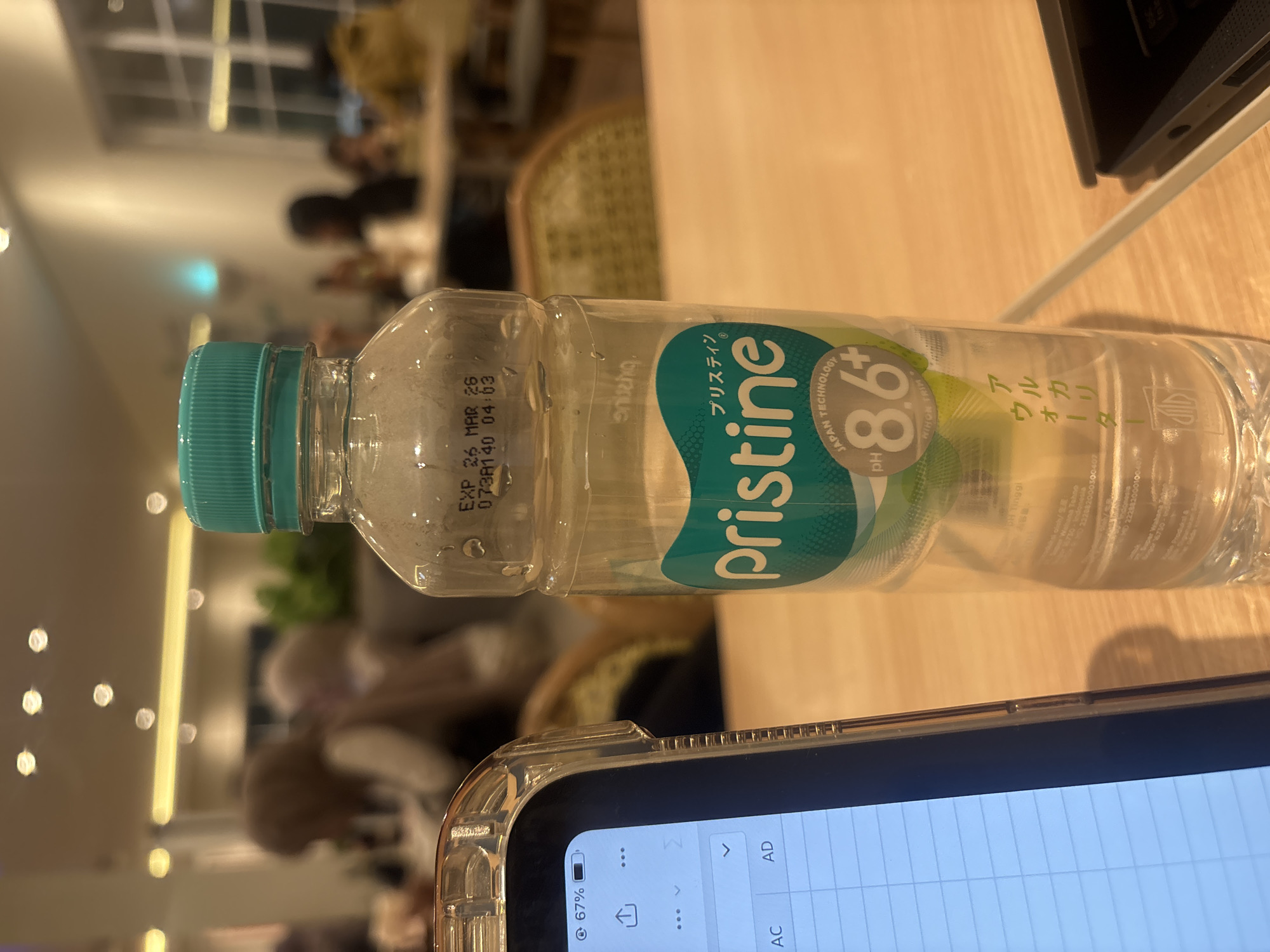 The image shows a bottle of Pristine Alkaline Water 8.6+, labeled in both Japanese and Indonesian, lying horizontally on a light wooden surface. The expiration date printed on the bottle reads "EXP 26 MAR 26". A portion of a tablet or smartphone, displaying a spreadsheet application, is visible in the foreground. In the blurred background, there are people seated in what appears to be a cafe or restaurant setting. Based on the label, the water is produced by PT Tirta Investama, a Danone company, in Indonesia. A more precise location cannot be determined from the provided image.