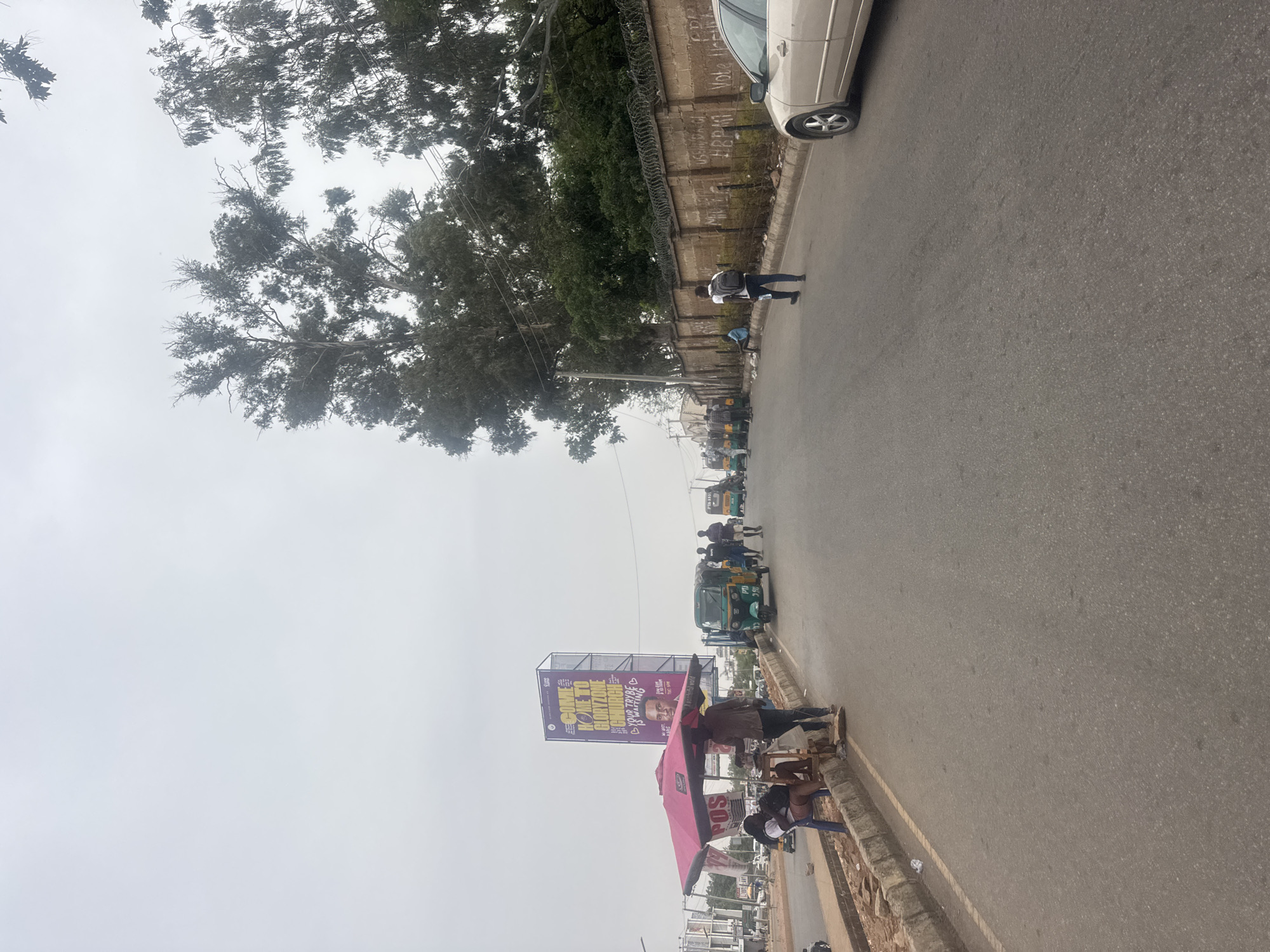 An overhead shot captures a street scene in Jos, Nigeria, characterized by an overcast or hazy sky. The asphalt road, running diagonally through the frame, is lined on one side by a curb and a wall topped with razor wire. Large, leafy trees grow behind this wall, their branches extending upwards and across the top of the image.
Several individuals are visible along the road. One person wearing a white shirt and black backpack walks on the paved area near the wall, while another is closer to a light-colored car partially visible at the top right. Further down the street, multiple green and yellow auto-rickshaws (Keke Napep) and more people indicate a busy section of the road. On the lower right side, a group of people congregates around a pink awning or stall.
A large billboard stands prominently in the mid-ground, displaying text that reads "COME HOME TO CHORIZONE" and "YOUR TRIBE IS WAITING," along with a portrait of a man and the phrase "MR MUST BE PRESENT TO VOTE" and "PITG.NET". Graffiti is scrawled on the wall, with phrases like "VOTE HRP" and "05 GANG" partially legible. The pink awning also shows "POS," suggesting a point-of-sale service. The scene depicts typical daytime street activity, with commuting and informal commercial interactions taking place.