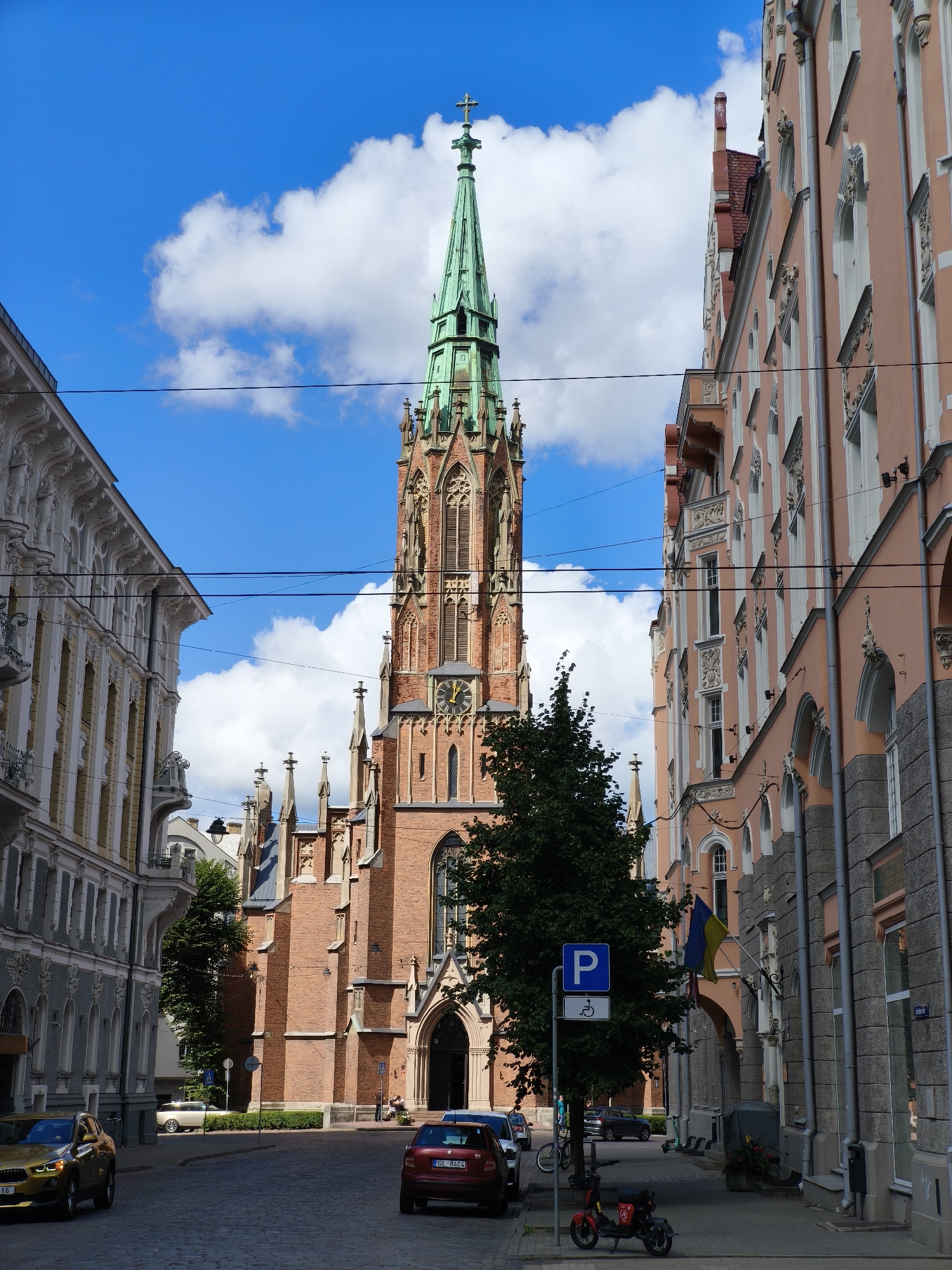 The image depicts a street view in Riga, Latvia, dominated by the St. Gertrude's Evangelical Lutheran Church. The church, constructed from red brick, features a prominent green spire topped with a cross. Flanking the church are two ornate buildings: one on the left with grey-toned facade and classical architectural elements, and another on the right with a salmon-pink facade and detailed decorative elements. The street is paved with cobblestones, and several cars are parked and driving along it. A blue parking sign with a wheelchair symbol is visible on the left side of the street, near a tree. The sky is bright blue with scattered white clouds. A Ukrainian flag is flying from a building on the right. There are no discernible people interacting.