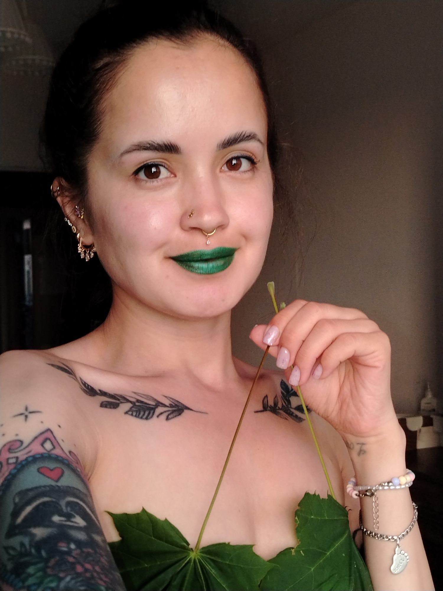 This is a head-and-shoulders portrait of a young woman taken indoors in Noviy, Russia, under natural light. The subject is a woman with dark hair pulled back, brown eyes, and light-toned skin. She is looking directly at the viewer with a slight smile, conveying a calm and confident expression.
She is wearing vibrant green lipstick. Her face features several piercings: multiple gold-toned earrings, including hoops with dangling gems and a purple stud in her left ear, a small gold stud on her left nostril, and a gold septum ring with a clear teardrop-shaped jewel.
Visible tattoos include a black leafy branch design stretching across her collarbone area, and a colorful sleeve tattoo on her left upper arm featuring a raccoon wearing a crown with a red heart on it, amidst other designs. Two large green leaves, possibly maple leaves, are strategically placed to cover her chest.
She is holding a thin green stem or twig in her right hand. Her fingernails are painted with a light, iridescent pink polish. On her right wrist, she wears two bracelets: one multicolored beaded bracelet and a silver chain bracelet with a footprint-shaped charm. The text "Таша" is clearly visible on the footprint charm. The background is dimly lit and nondescript, appearing dark gray or brown, suggesting an indoor setting with possibly a window to the left providing the light source.