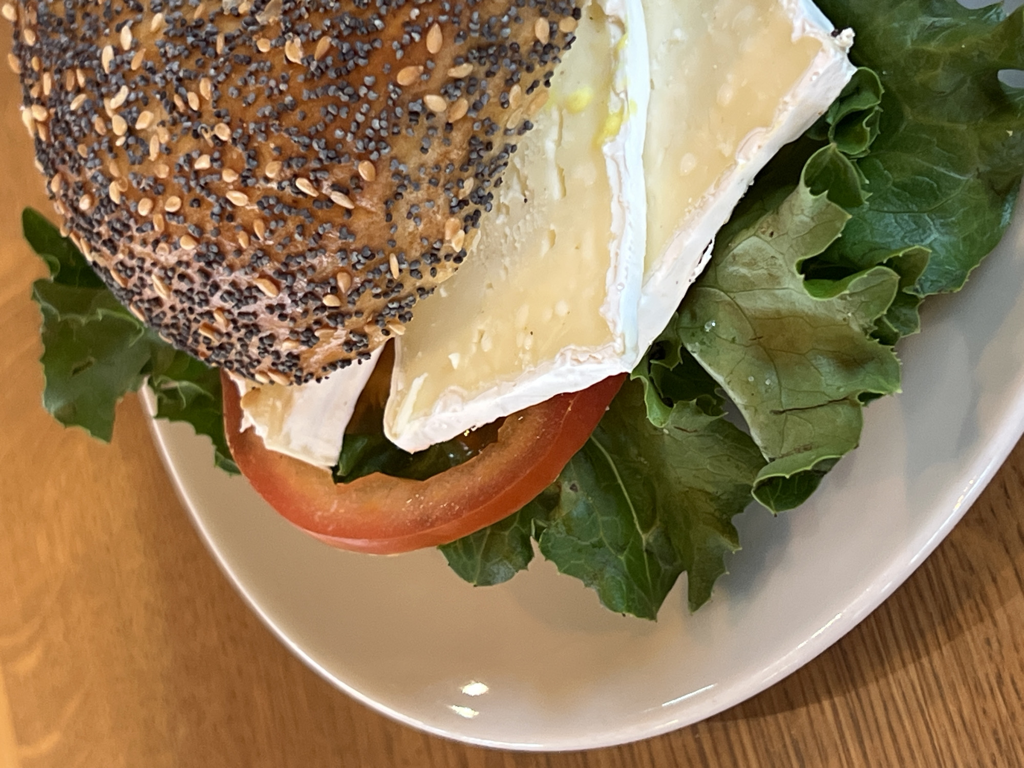 The image displays a close-up, overhead view of a sandwich on a white plate, positioned on a wooden surface. The sandwich features a poppy seed and sesame seed bagel, sliced in half, with the top half visible. Between the bagel halves are layers of green lettuce, a slice of red tomato, and multiple slices of white cheese. The cheese appears to be a soft, creamy variety, possibly brie or camembert, with some yellowing indicating ripeness. The lighting suggests it is indoors, possibly a restaurant or cafe setting, with a natural light source illuminating the food from the side. There is no visible indication of the city of Moosburg, Germany, or any specific location within it.