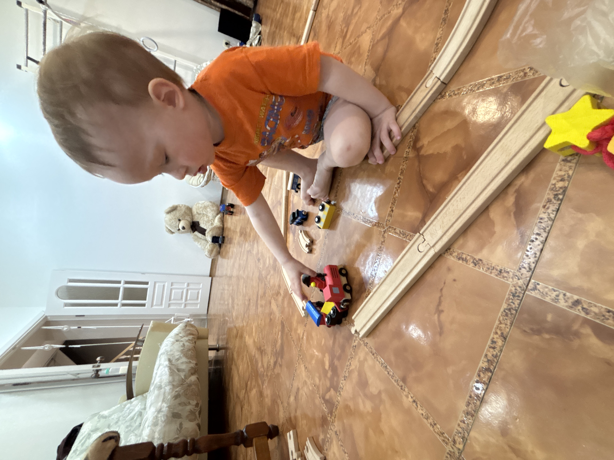 A young child in an orange t-shirt is sitting on a brown tiled floor, playing with wooden toy trains and tracks. A teddy bear sits near a white door in the background. The location is likely indoors, potentially a residence. There is no evidence that this image is in Ferma, Russia.