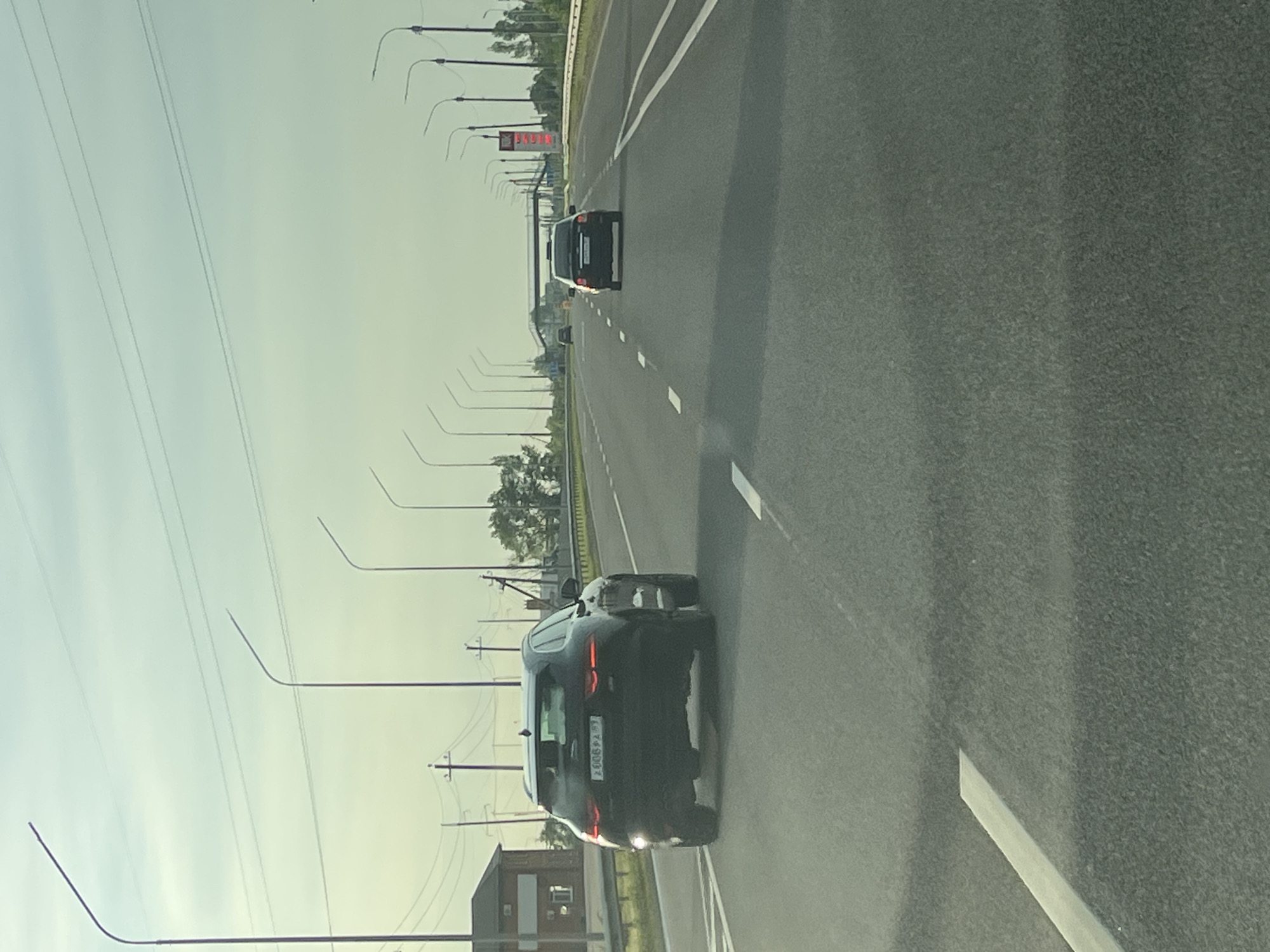 The image depicts a highway scene in the city of Psekups, Russia, under an overcast sky. The viewpoint is from a vehicle traveling on the highway.
In the foreground, a black Jaguar SUV is prominently featured in the right lane, moving away from the viewer. Its license plate, which appears to be from the Krasnodar Krai region of Russia, is partially visible and reads "E 606 PA 93". The car's taillights are illuminated.
Further down the highway, in the same lane, a dark-colored sedan is visible, also traveling away from the viewer. Beyond that, an overpass crosses the highway, with a pedestrian walkway above.
To the left of the highway, a building with a red roof and brick facade is visible, along with utility poles and power lines. A variety of streetlights line both sides of the highway, stretching into the distance.
The highway itself is paved with asphalt and marked with white lane dividers. The overall environment suggests a typical highway setting on a cloudy day. There are no people visible in the frame, and the focus is on the vehicles and infrastructure.