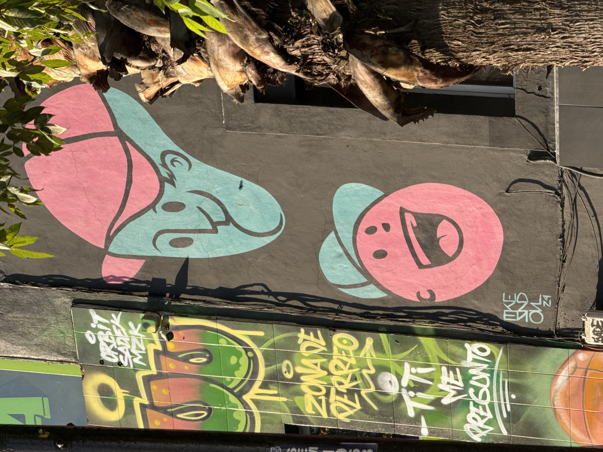 A mural in Puebla, Mexico, featuring pink and blue cartoonish figures on a gray wall beneath a tree trunk. Below the mural, a graffiti panel displays green, yellow, and orange designs with white lettering. Wires and utility boxes are visible on the right side of the gray wall.