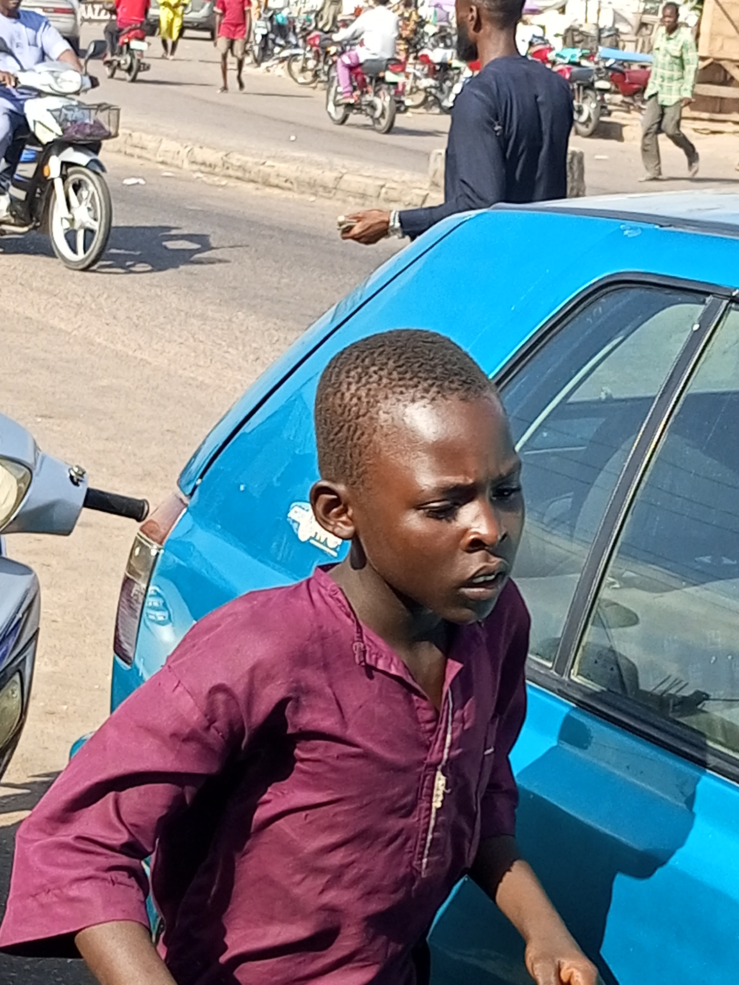 In Gombe, Nigeria, on a sunlit, dusty street, a young boy with dark skin and short hair, wearing a deep magenta tunic, stands next to the passenger side of a bright blue car. His gaze is directed forward and slightly to the right, with a neutral expression. The car's window is down, revealing the interior and a faint reflection of objects. To the left of the boy, the front of a light-colored motorcycle is partially visible, with its handlebars and a front basket. In the background, the street is active with traffic. A man in a dark blue garment walks away from the camera. Further in the background, motorcycles and pedestrians are moving, suggesting a busy urban environment. The scene is typical of a street in Gombe, characterized by its vibrant colors, bustling activity, and the presence of common modes of transportation like motorcycles.