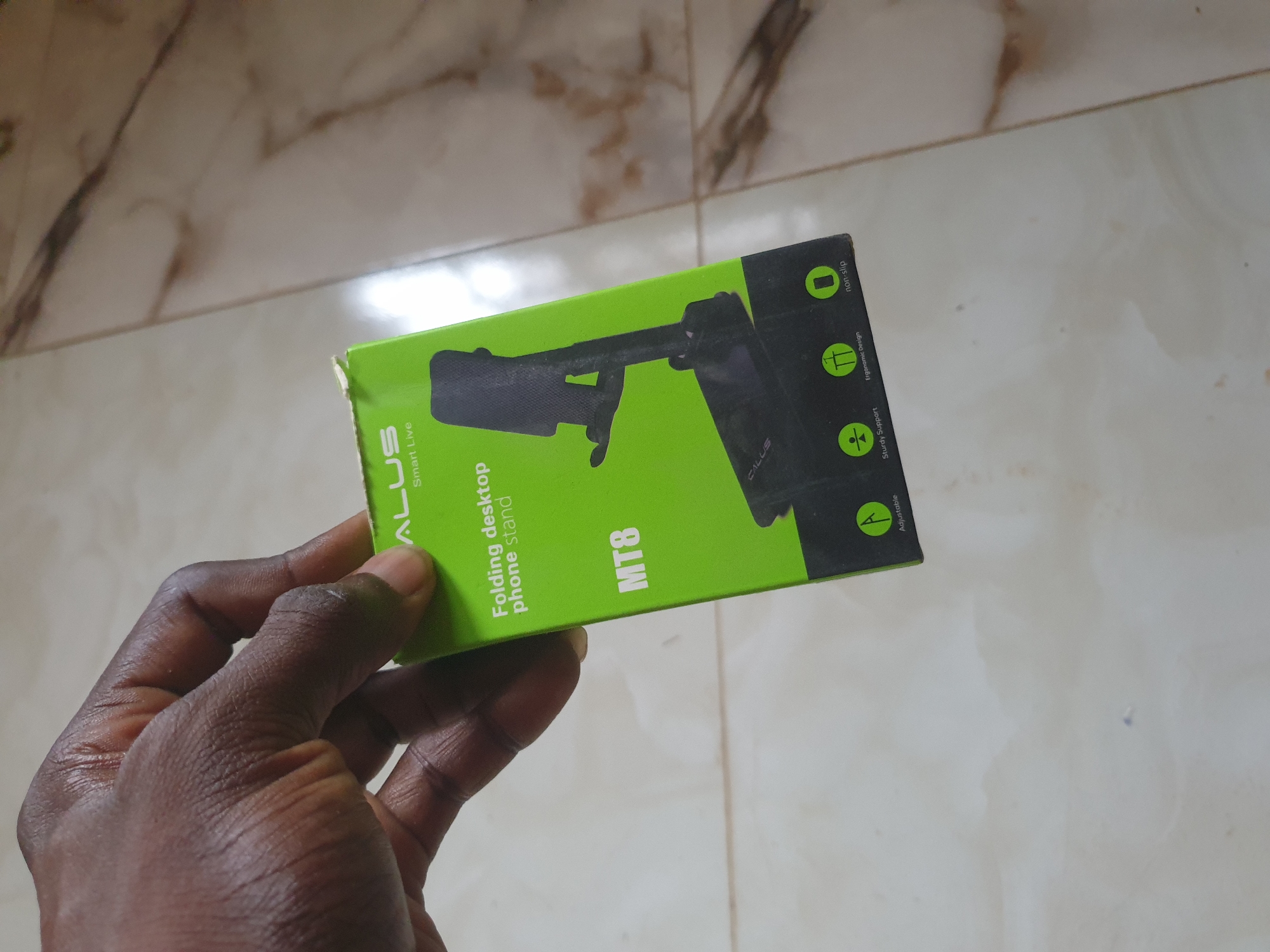 A dark-skinned hand is holding a green and black box for a folding desktop phone stand. The box has text and icons on it. The visible text on the box includes "ALUS Smart Live", "Folding desktop phone stand", "MT8", "Non-slip", "Ergonomic Design", "Sturdy Support", and "Adjustable". The background is a tiled floor with a marble pattern. The lighting appears to be natural daylight. The image was taken in Anilelerin, Nigeria.