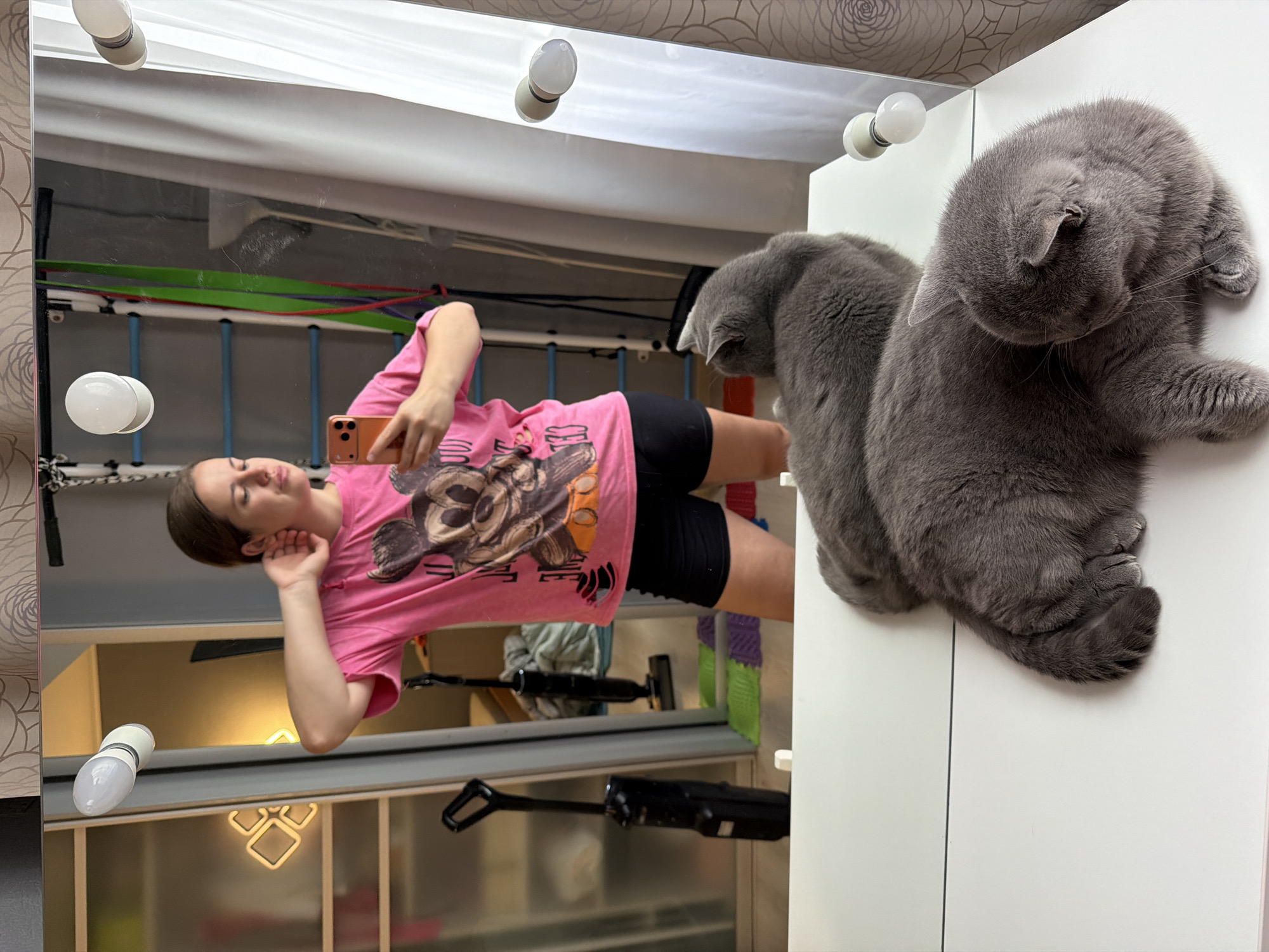 A mirror reflects a woman taking a photo of herself with a mobile phone, and two gray cats are perched on the white cabinet to the right of the mirror. The mirror is framed with lightbulbs. Behind the woman, reflected in the mirror, are colorful resistance bands and what appears to be a weight rack. The background includes a glimpse of another room with a modern chandelier. The location in Sochi, Russia, cannot be determined from the image content.