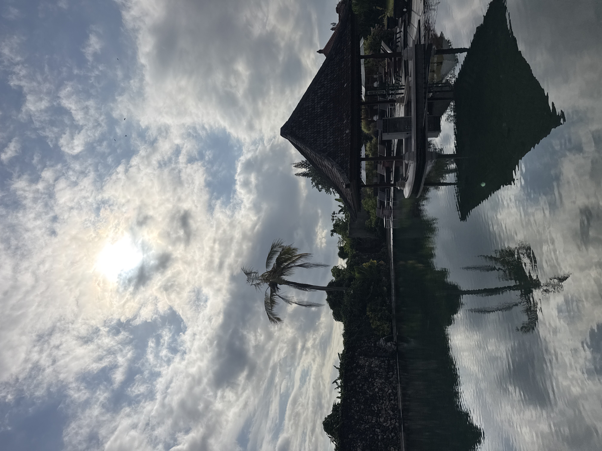The image, likely taken in Kuta Selatan, Indonesia, shows a scenic view with reflections. The sky is filled with clouds and a bright sun peeks through. A palm tree stands tall amidst lush greenery. There is a building with a tiled roof, possibly a gazebo or outdoor structure. Reflections of the sky, clouds, building, and trees are visible in the still water, creating a mirrored effect.