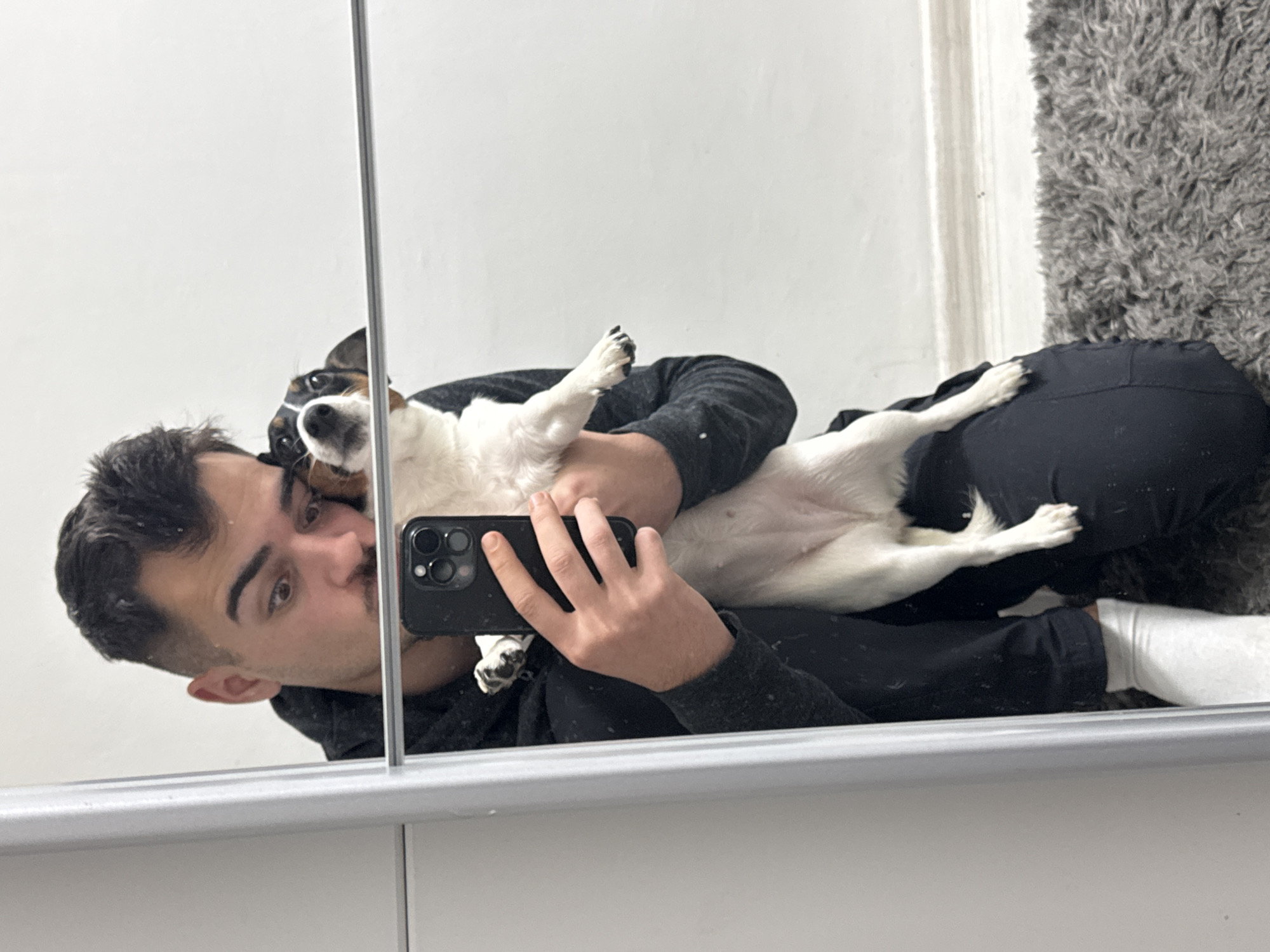 A mirror selfie captures a man lying on his back, holding a small dog on his chest in what appears to be an indoor setting in Sofia, Bulgaria. The man, with dark hair, a short beard, and wearing a dark hoodie and sweatpants with white socks, holds a black smartphone to take the picture. His eyes are directed towards the camera with a neutral to slightly amused expression.
Cradled against him, the dog—a small, mostly white breed with brown and black patches on its head and back, possibly a Jack Russell Terrier—looks attentively towards the camera, resting its head near the man's shoulder. Its front paws are resting on the man's chest.
The scene is reflected in a large mirror, which extends from a lower white cabinet or surface upwards. To the right of the man and dog, a textured, dark gray shaggy rug is visible, suggesting a cozy, domestic environment. The overall lighting is bright and even, indicating either ample artificial light or natural light from an unseen source. The perspective of the photo makes the subjects appear to be lying horizontally. No specific time of day or weather can be discerned, and there is no visible text.
