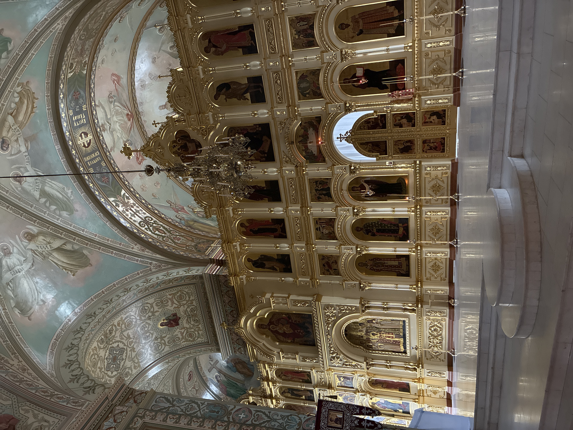 This is an interior view of the Church of the Intercession on the Nerl, located in Боголюбово, Russia. The image captures the intricate and ornate architecture of the church, focusing on the ceiling and walls. The ceiling is decorated with religious paintings and intricate patterns. A large, ornate chandelier hangs from the ceiling, adding to the grandeur. The walls feature a multi-tiered iconostasis with numerous religious icons and gold detailing. The overall environment is rich in religious symbolism and historical significance. The floor, partially visible, is made of light-colored stone or tile. The lighting is soft, enhancing the delicate details of the artwork and architecture. The image is taken from a low angle, emphasizing the height and majesty of the church interior.