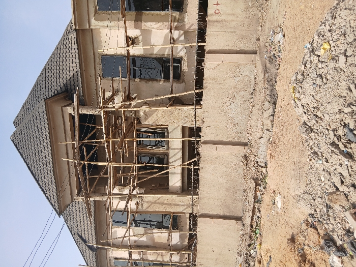 The scene depicts a two-story residential building under construction in Mgbidi, Nigeria. The structure features a multi-faceted, dark grey tiled roof system and unpainted, textured exterior walls. Extensive scaffolding, constructed from wooden poles and planks, is erected across the entire front facade of the building. Multiple dark-paned windows are visible across both floors, some appearing installed. In the immediate foreground, a concrete perimeter fence separates the building from a dirt ground strewn with construction debris, loose soil, and scattered refuse including plastic bags and wrappers. No individuals are present. Overhead, utility lines traverse a clear, light blue sky.