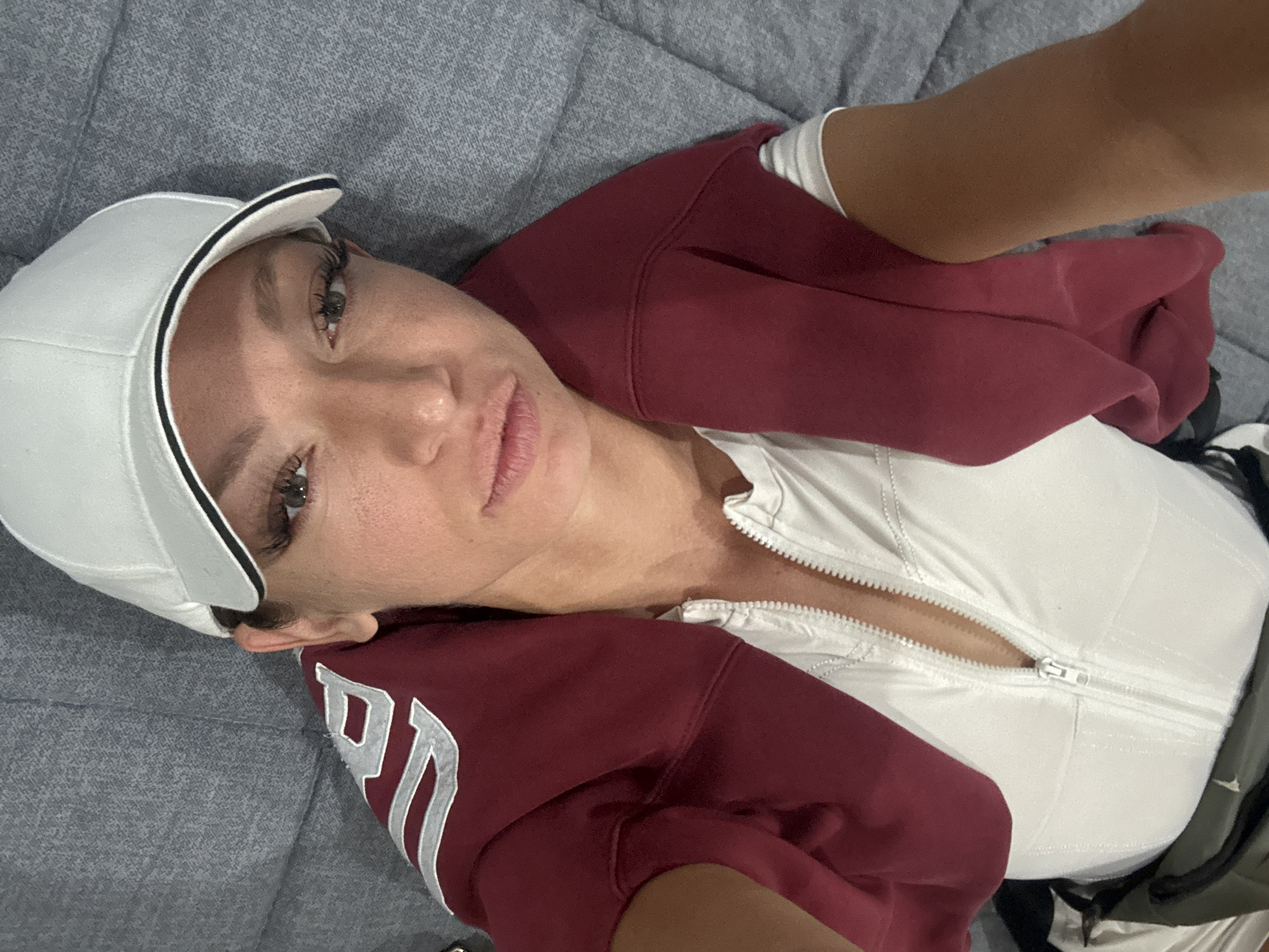 A woman wearing a white baseball cap and a partially unzipped white top with a maroon sweatshirt around her shoulders is lying on a gray quilted surface, possibly a bed or couch. The sweatshirt appears to have the letter "N" or "P" in white on its sleeve. The woman is looking directly at the camera. The environment is indistinct, giving no evidence of a specific location in Baan Chaweng, Thailand.