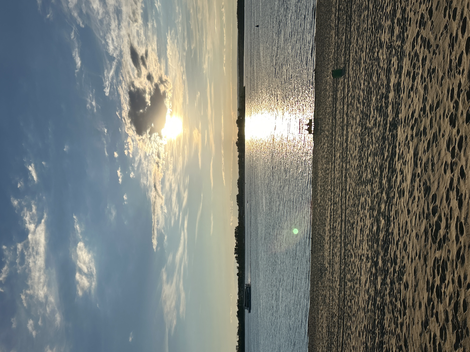 This image depicts a serene beach scene at sunset in Novokuybyshevsk, Russia. The sun, low in the sky, casts a brilliant, shimmering reflection across the calm water. Wispy clouds are scattered across a pale blue sky, illuminated by the golden light of the setting sun.
The foreground is dominated by the textured surface of the sandy beach, with its characteristic patterns of footprints and shadows. To the left, the gentle waves of the water meet the shore, creating a tranquil boundary. A distant shoreline is visible on the horizon, lined with trees.
In the water, a small boat or barge can be seen moving slowly. Closer to the shore, a pair of figures are seated on what appears to be a bench or a low structure, silhouetted against the bright glare of the sun on the water. A small green object, possibly a marker or a piece of equipment, stands on the sand near the water's edge.
The overall atmosphere is peaceful and tranquil, characteristic of a late summer evening. The warm hues of the sunset evoke a sense of calm and relaxation. There is no visible text in the image.
