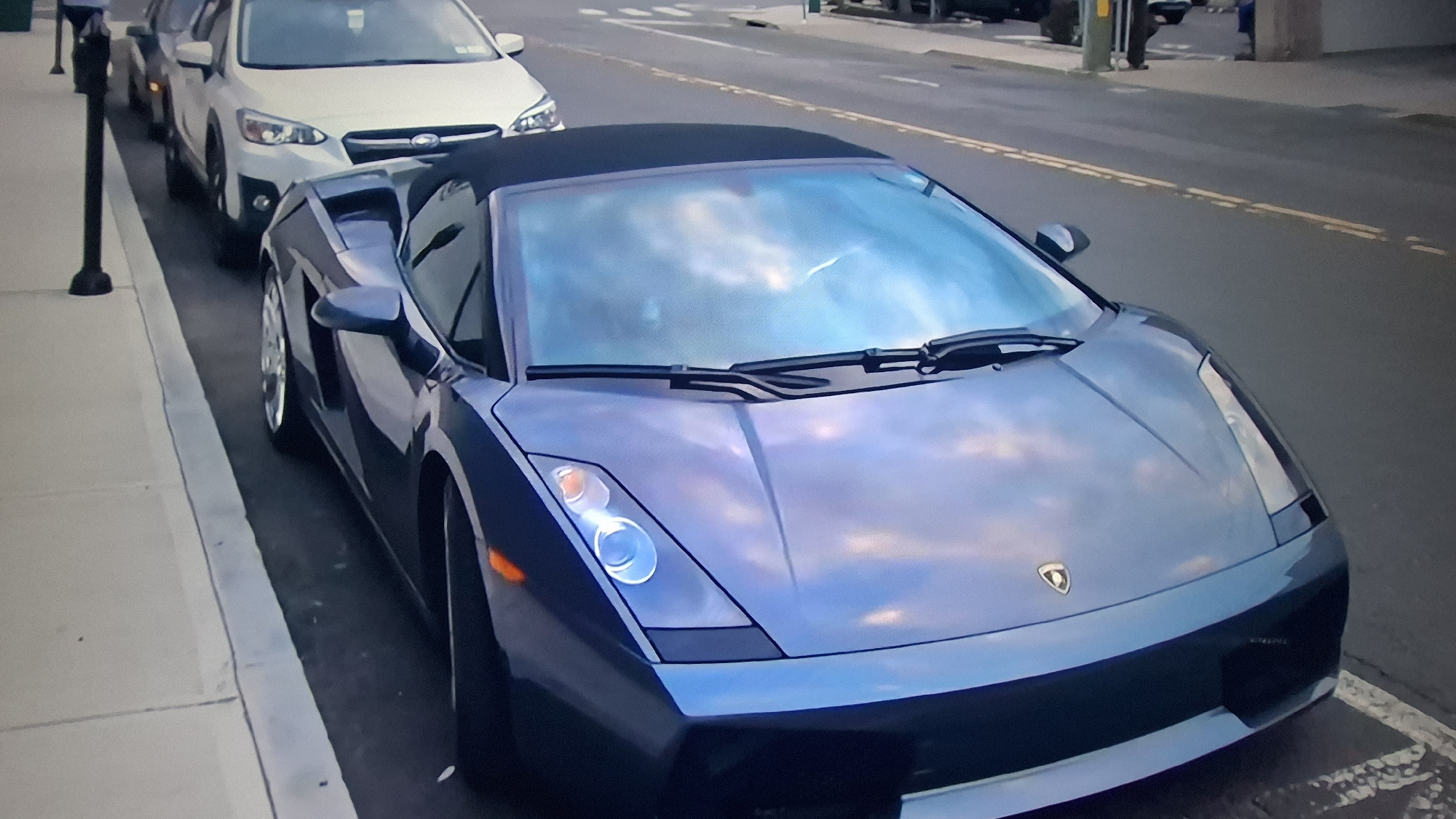 An eye-level, slightly downward-angled shot captures a metallic blue or grey Lamborghini Gallardo Spyder convertible parked at the curb of what appears to be an urban or suburban street. The car is the primary subject, occupying the foreground and center-right of the image. Its sleek design and a soft, dark fabric convertible top are visible. The windshield and hood brightly reflect a light blue, cloudy sky, suggesting daytime conditions with either scattered clouds or an overcast sky.
Behind the Lamborghini, a white Subaru Crosstrek is parked, followed by another dark-colored vehicle further down the street. To the left of the parked cars, a clean, light-colored sidewalk is visible, with a dark, slender pole (possibly a lamppost or parking meter pole) at its edge in the far left background. To the right of the Lamborghini, the asphalt street features prominent double yellow lines, indicating a no-passing zone. The front of the Lamborghini displays its distinctive emblem, a rearing bull, centered on the hood. No people are clearly visible in the frame, and there is no discernible activity other than parked vehicles. The scene depicts a quiet street setting.