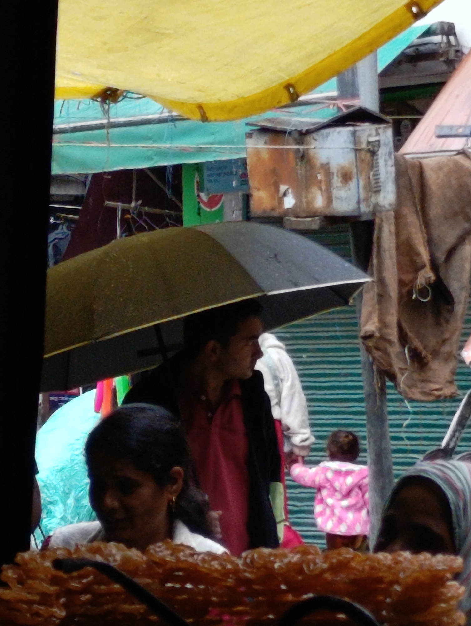 The image depicts a street scene in Manali, India, under an overcast sky, suggesting it may be raining or recently rained. A large yellow tarp is suspended overhead, partially obscuring the view of the sky. In the foreground, a woman with dark hair and wearing a light-colored top is partially visible, looking down. In front of her, a pile of fried, golden-brown sweets, possibly jalebi, is arranged in a lattice-like pattern.
Behind the woman, a man holding a black and grey umbrella stands partially obscured. He is wearing a dark jacket over a pink shirt. To his left, another umbrella, this one light blue, is visible. Further back, a young child in a pink jacket with white polka dots is being led by an adult, though the adult's face is not clearly visible. The background features a green striped wall or structure, and various stalls or shop fronts with hanging fabrics and weathered metal structures. The overall atmosphere is that of a busy marketplace.