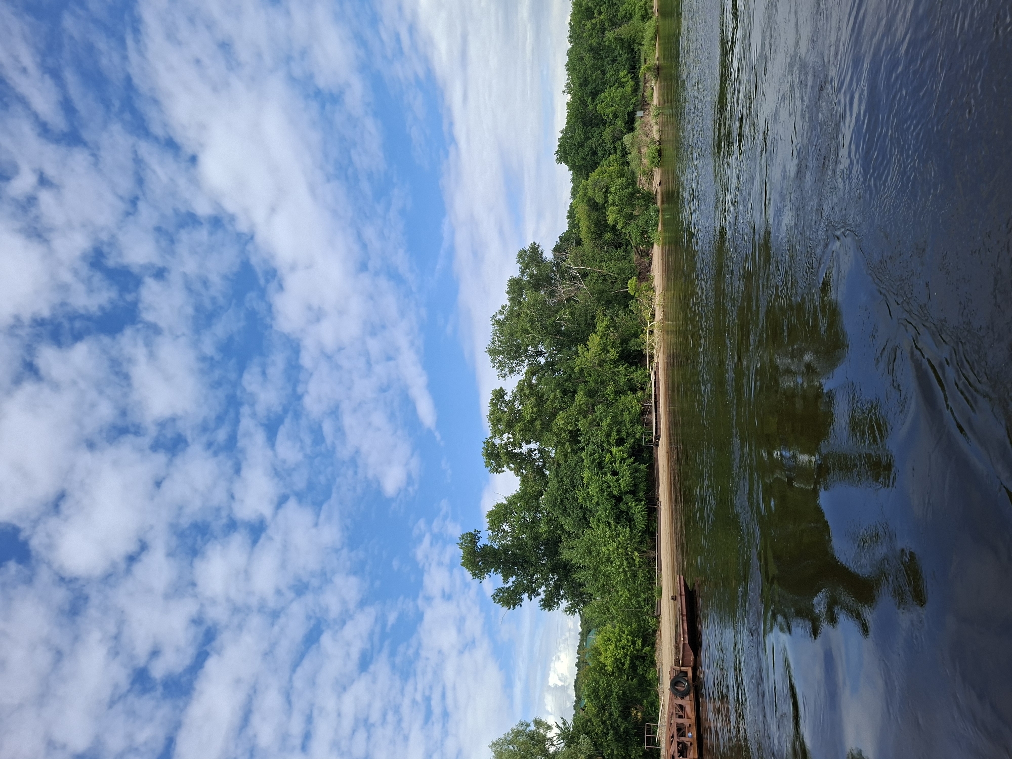 This is a wide-angle, eye-level, outdoor shot of a riverbank on a partly cloudy day. The river is dark and reflective, with ripples on the surface. On the left, a rusty barge is docked. The shore is sandy, with a line of dense green trees and foliage behind it. The trees vary in height and density, forming a lush backdrop. Above the trees, a bright blue sky is dotted with white, puffy clouds. The scene appears tranquil and natural, likely captured in a rural or park-like setting within or near Kazan, Russia, possibly along the Volga River.